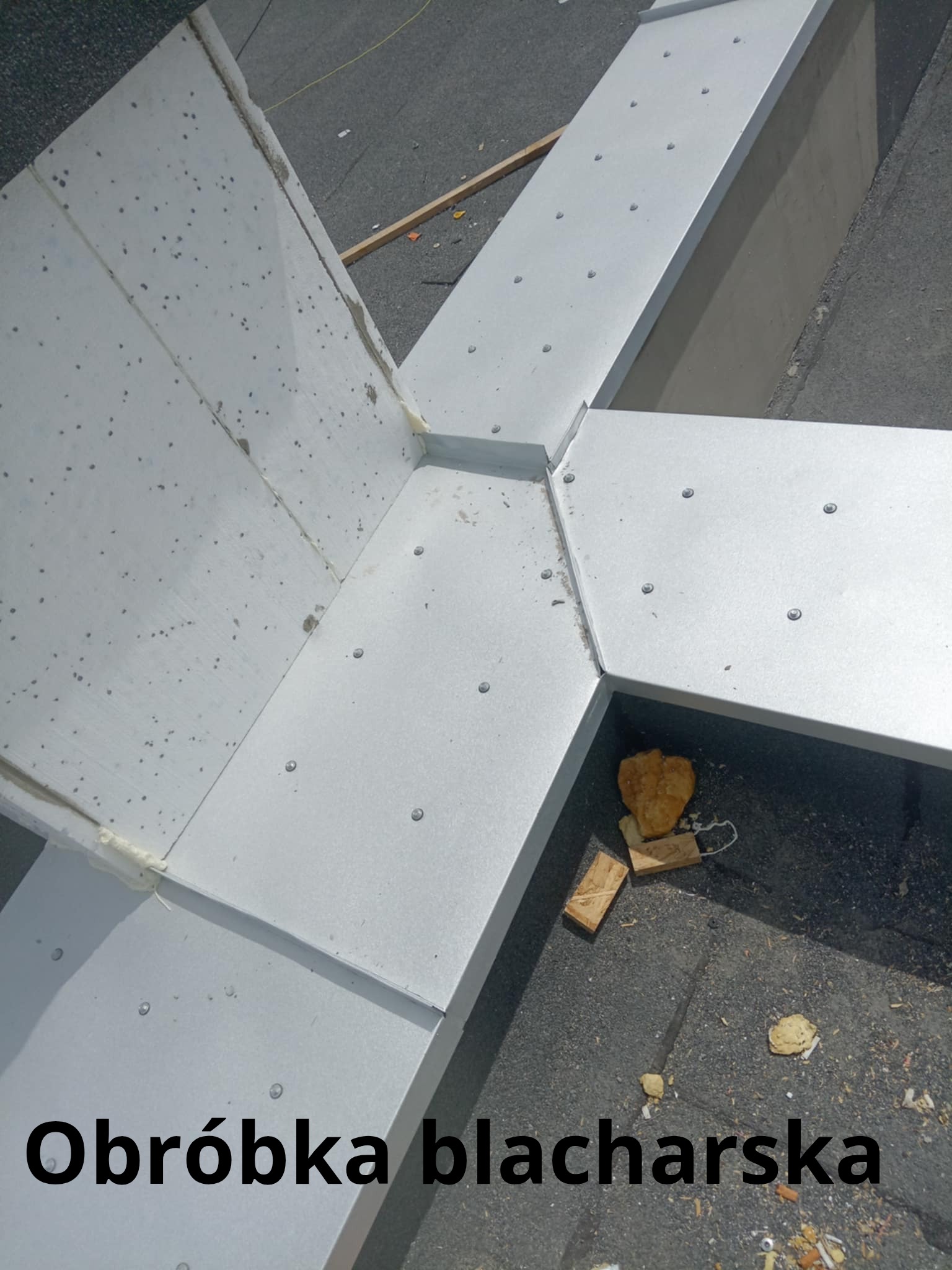 Szczegółowe ujęcie nowej, srebrnej obróbki blacharskiej dachu z widocznymi nitami i szczelinami wypełnionymi uszczelniaczem. Fragmenty gruzu na czarnym dachu.