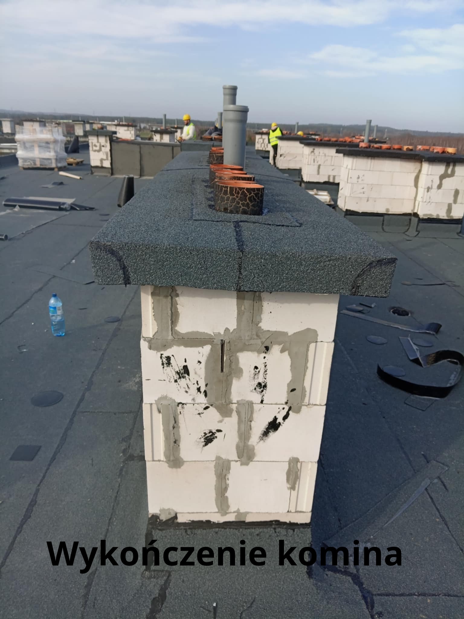 Wykończony komin na dachu pokrytym papą, z widocznymi obróbkami blacharskimi i pracownikami w oddali, podczas słonecznego dnia.