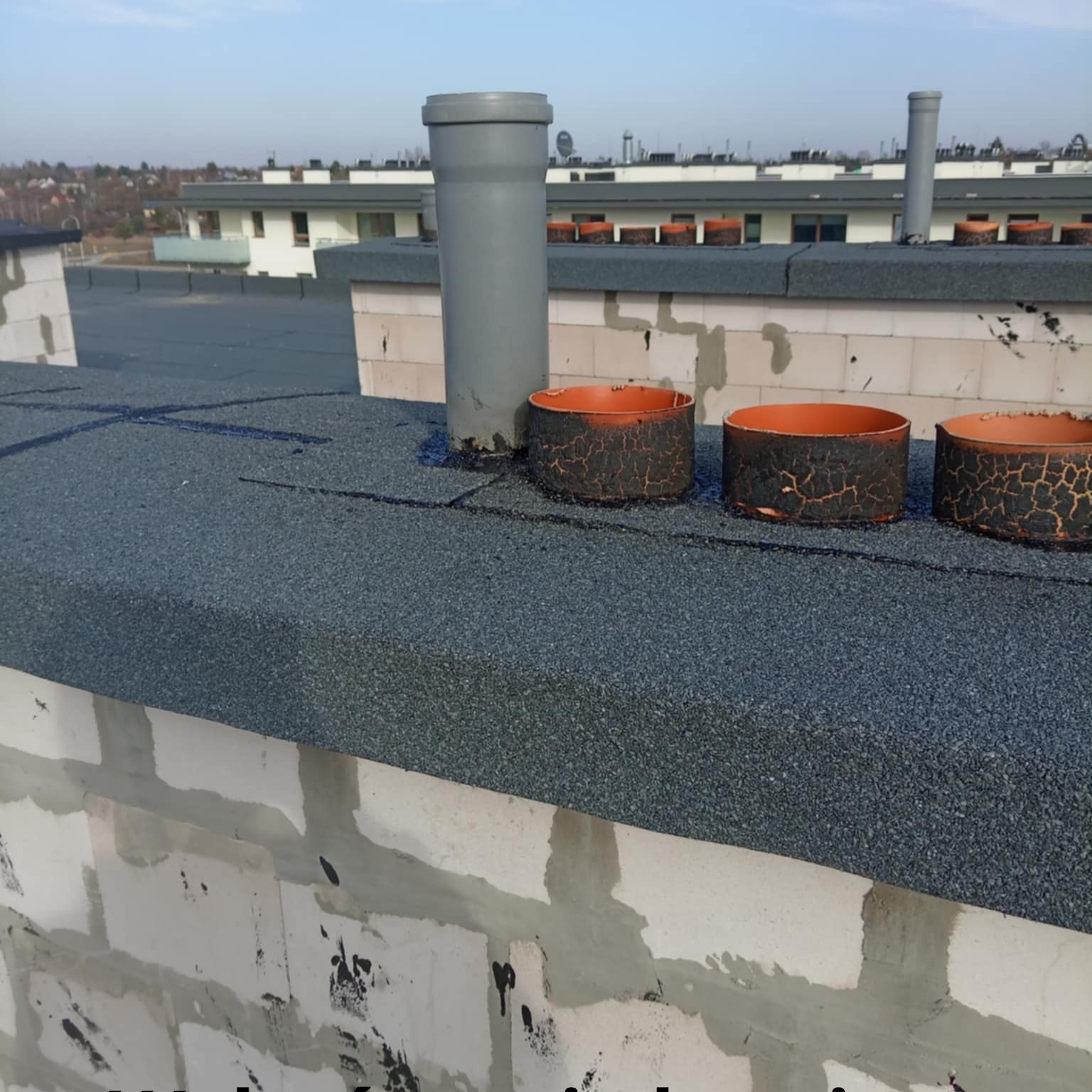 Dach pokryty papą z kominami wentylacyjnymi i obróbkami blacharskimi, widok z góry na tle zabudowy miejskiej w słoneczny dzień.