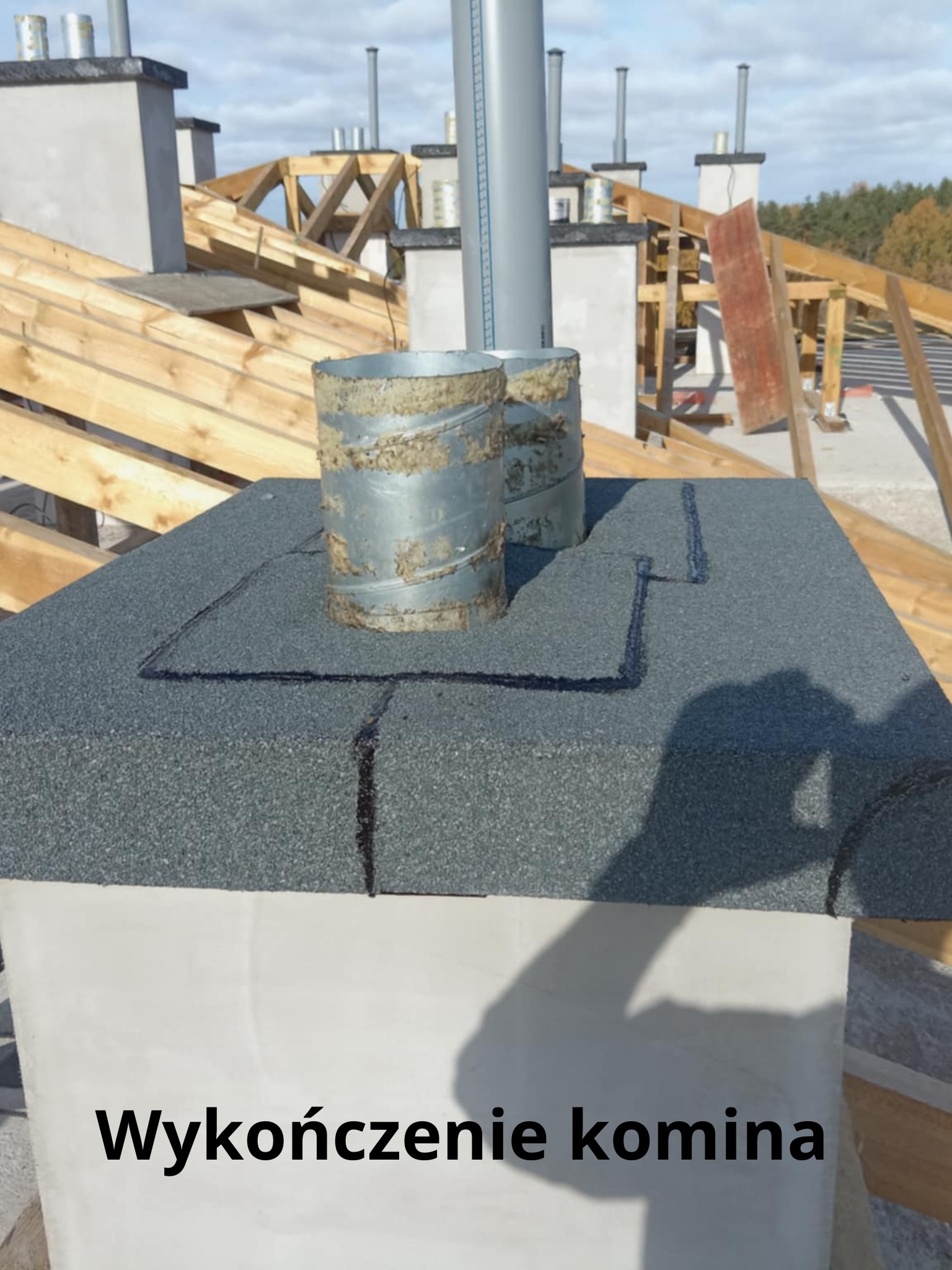 Szczegółowe wykończenie komina na dachu z papy, obróbka blacharska wokół komina, widoczne elementy konstrukcji dachu i inne kominy w tle.