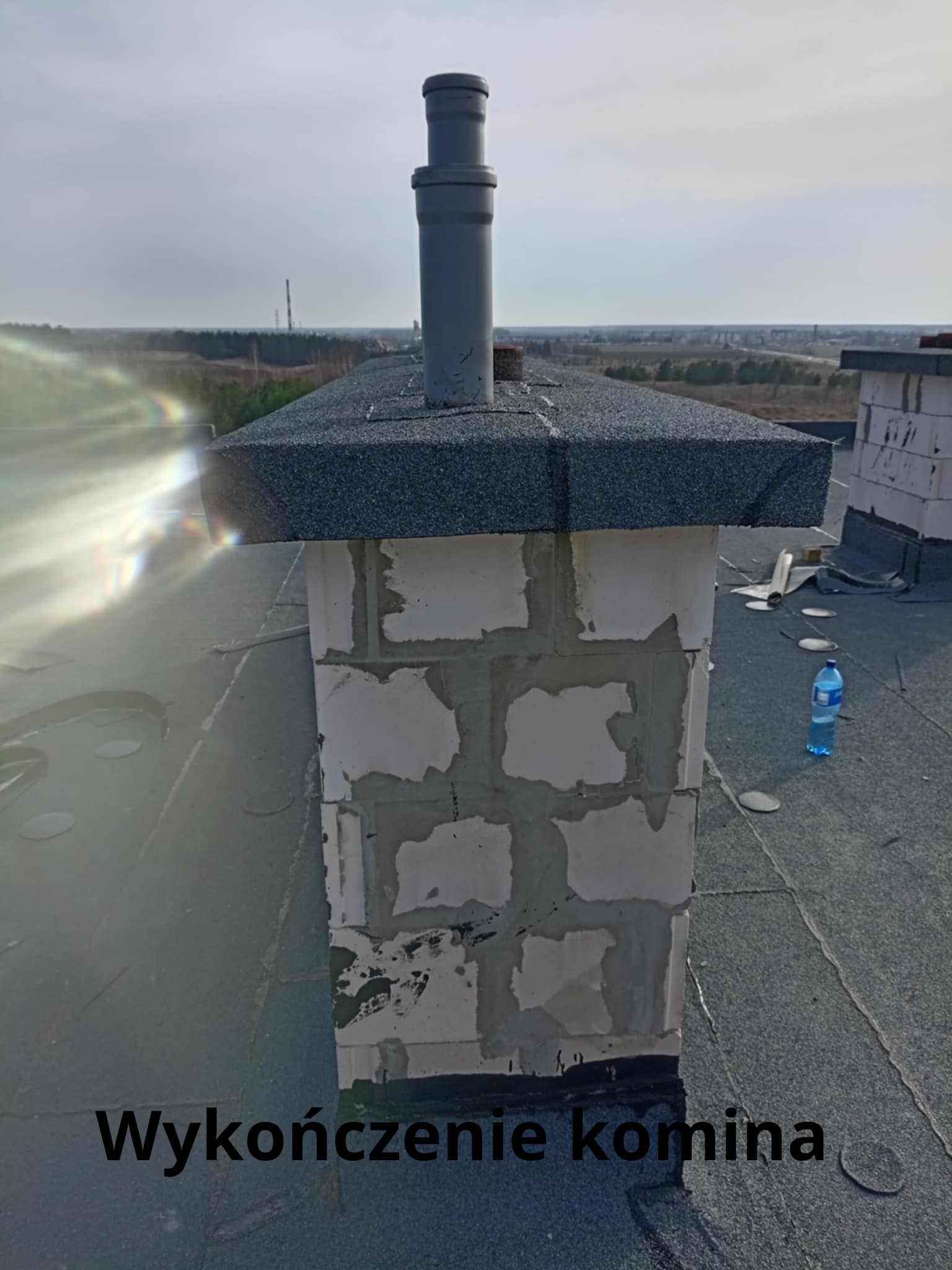 Wykończony komin z szarym daszkiem na dachu pokrytym papą. Widoczne bloczki kominowe i plastikowa rura. Butelka wody na dachu. W tle las i zabudowania.