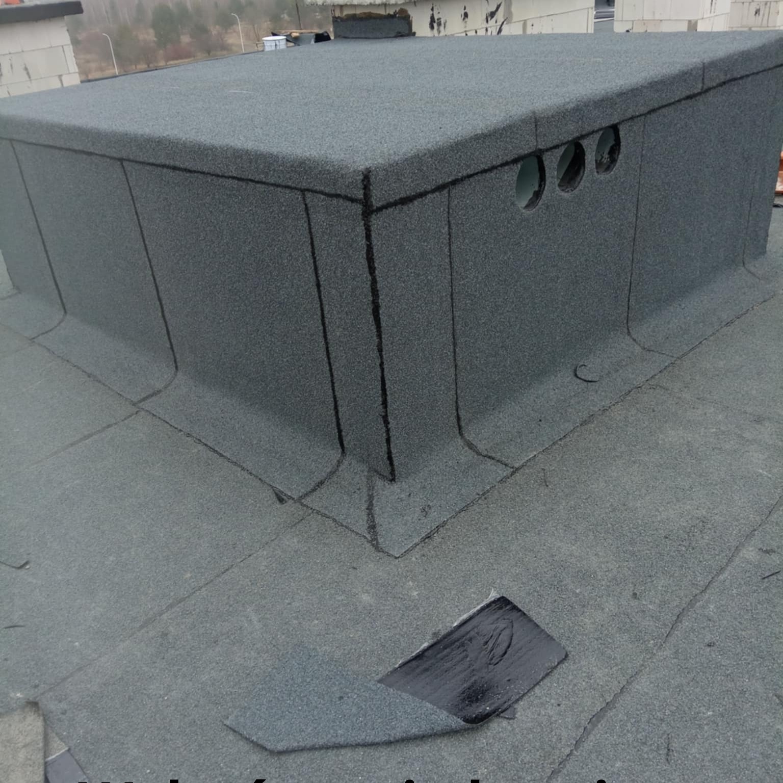 Szczegółowe ujęcie płaskiego, pokrytego papą dachu, z widocznymi detalami obróbek i komina wentylacyjnego. Fragment odklejonej papy na pierwszym planie.