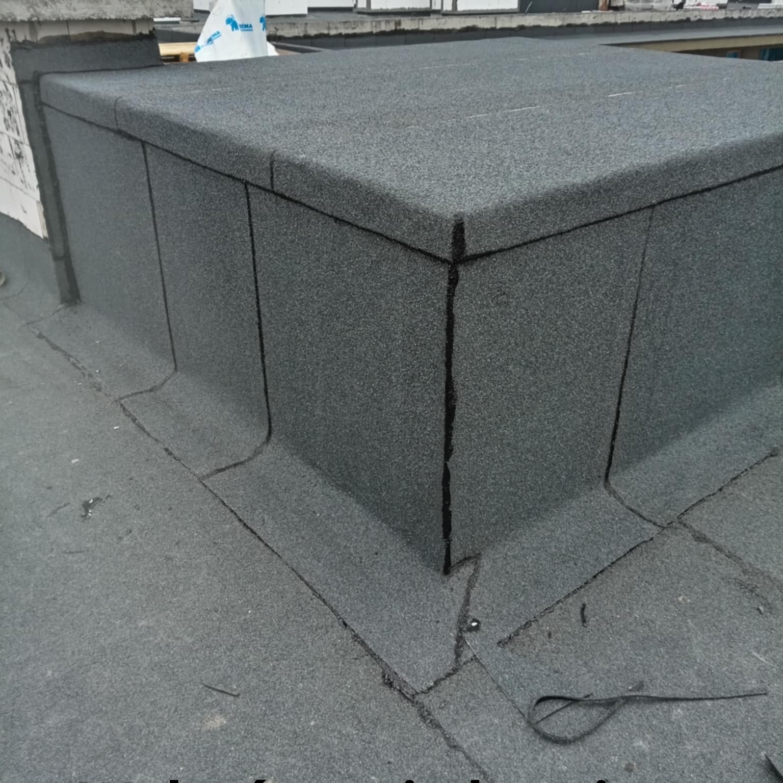 Szczegółowe ujęcie hydroizolacji dachu płaskiego z papy termozgrzewalnej, ukazujące precyzyjne łączenia i obróbki blacharskie komina. Widoczne detale wykonania.