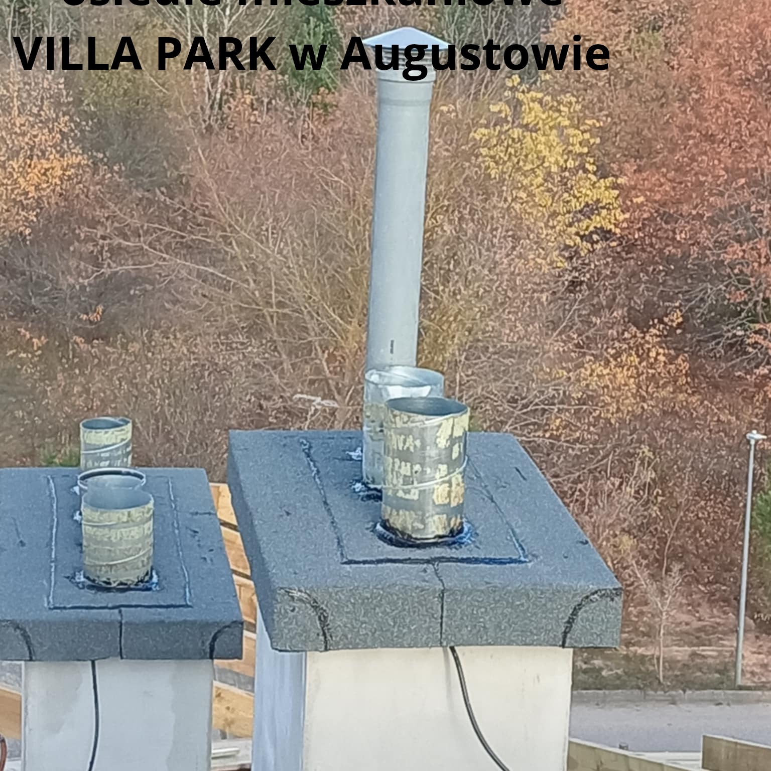 Szczegółowe ujęcie kominów z obróbkami blacharskimi na dachu budynku z widokiem na jesienne drzewa w tle. Widoczne elementy uszczelniające i rury wentylacyjne.