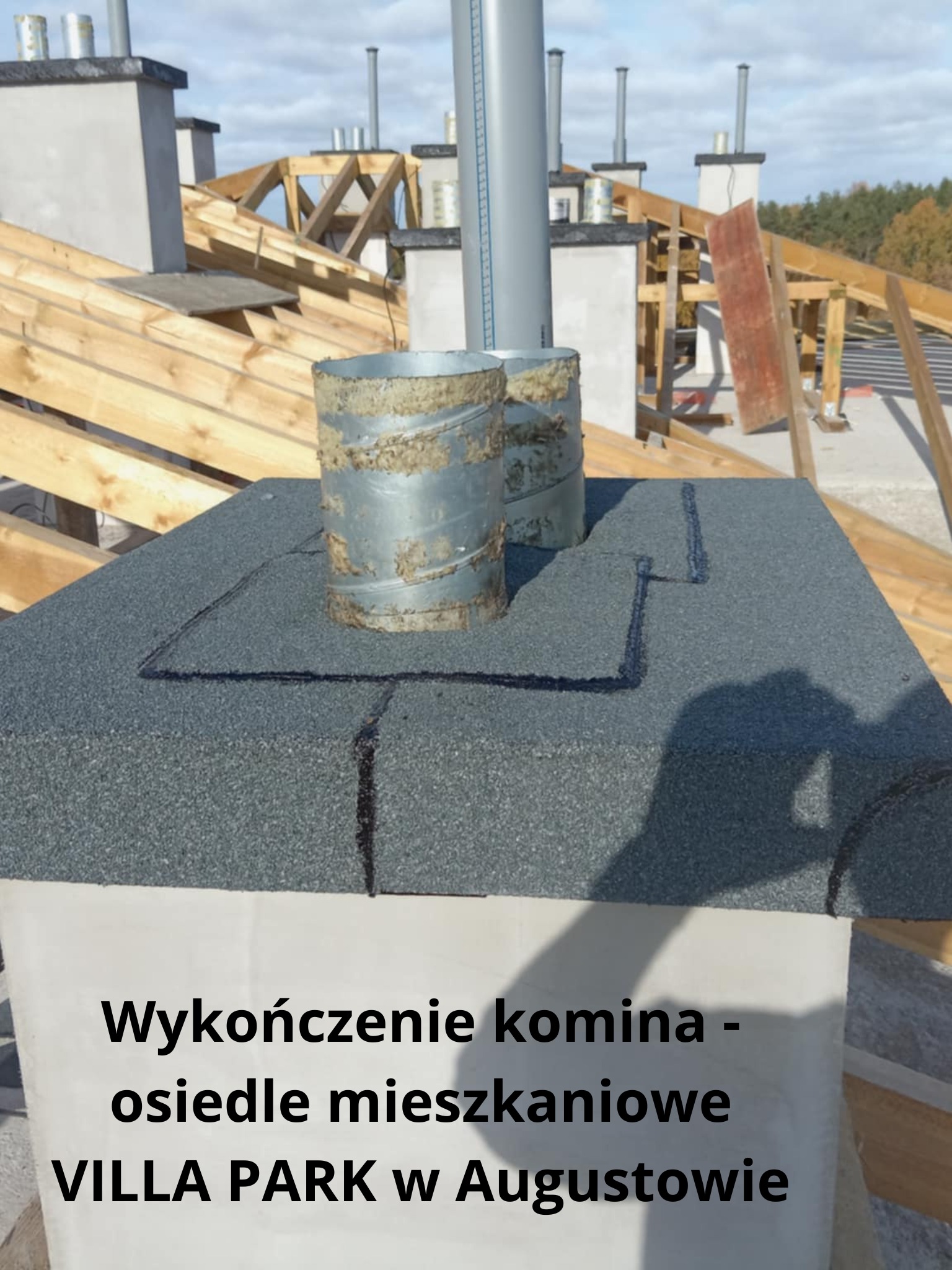 Wykończony komin na dachu z papy, z widocznymi obróbkami blacharskimi i elementami konstrukcji dachu w tle, na tle błękitnego nieba.