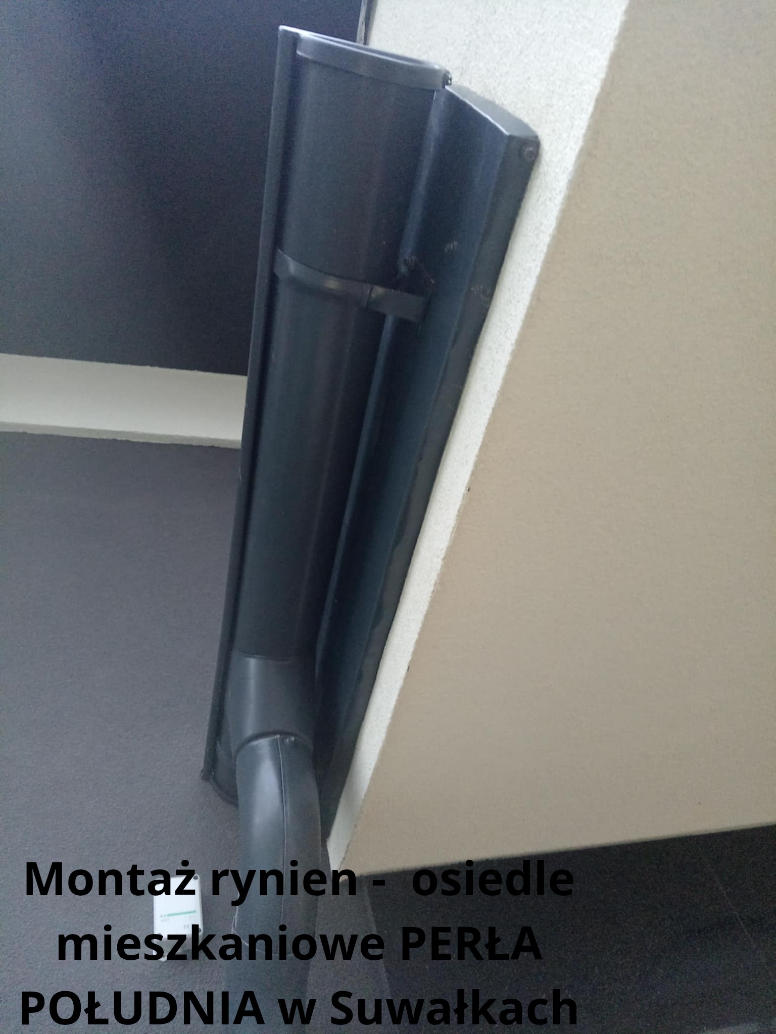Zamontowana, czarna rynna pionowa przy jasnej ścianie budynku mieszkalnego, widok z dołu, osiedle Perła Południa w Suwałkach.