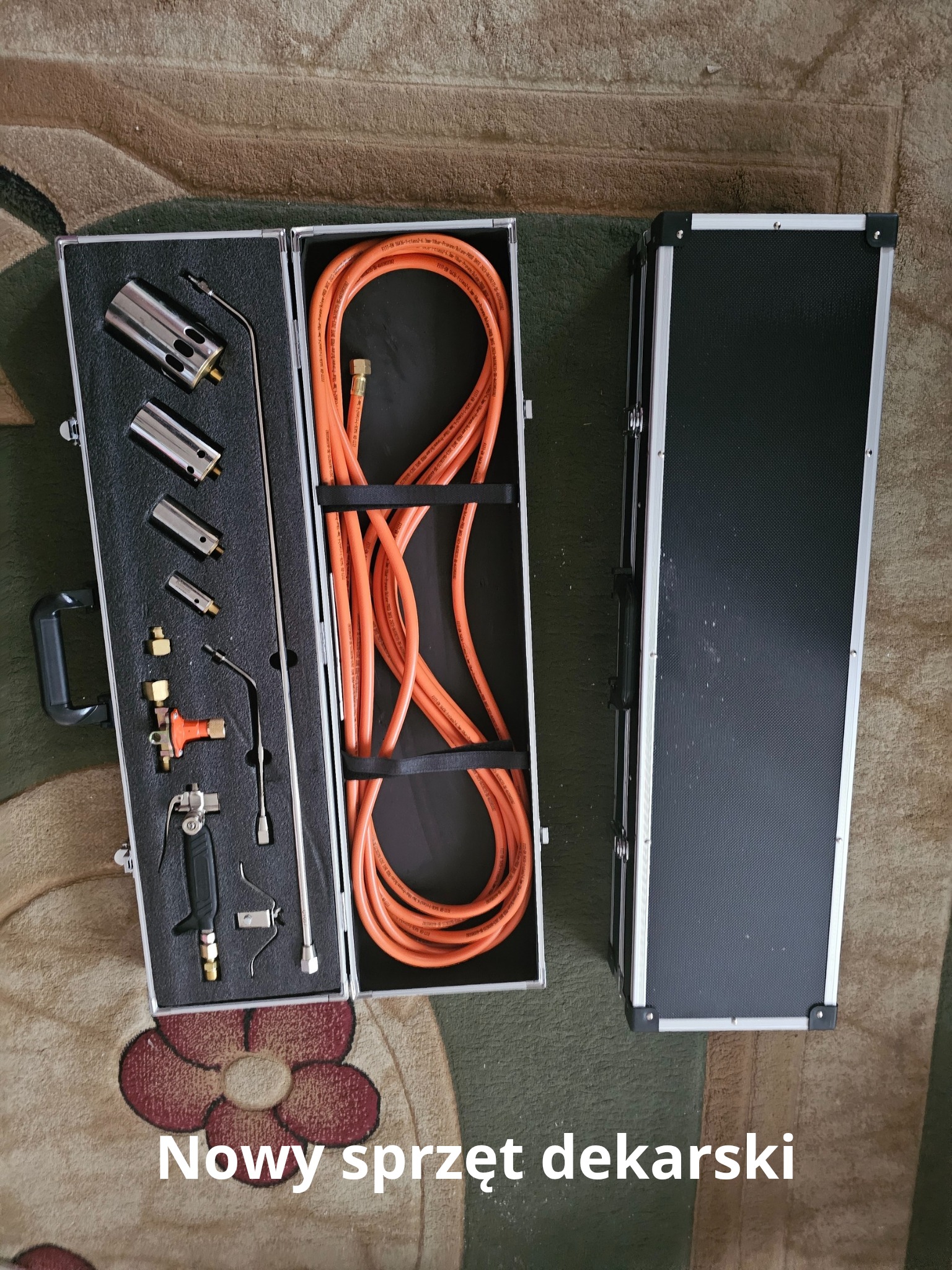 Otwarta walizka z kompletnym zestawem narzędzi dekarskich, w tym palnik i węże, ułożonych na wyściełanym wnętrzu, obok zamkniętej walizki.