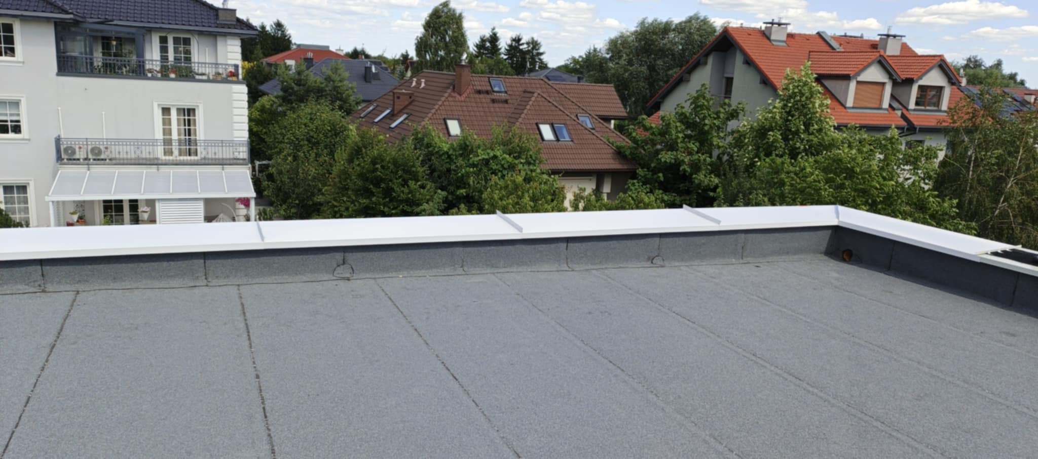 Szary dach płaski z białym wykończeniem na tle zabudowy miejskiej i zieleni. Widok z góry ukazuje detale pokrycia i otoczenie.