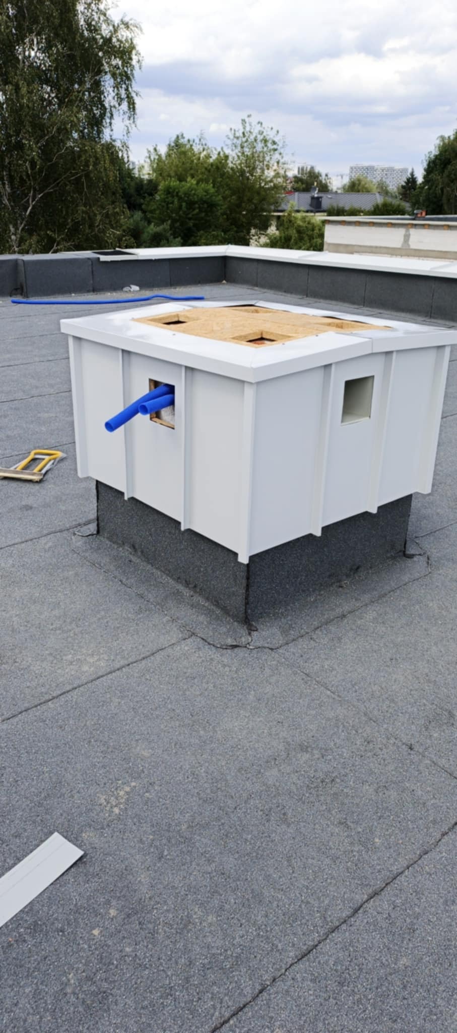 Biała obudowa komina wentylacyjnego na dachu pokrytym papą, z widocznymi niebieskimi rurami i drewnianym wykończeniem górnej części. Na dachu leżą narzędzia.
