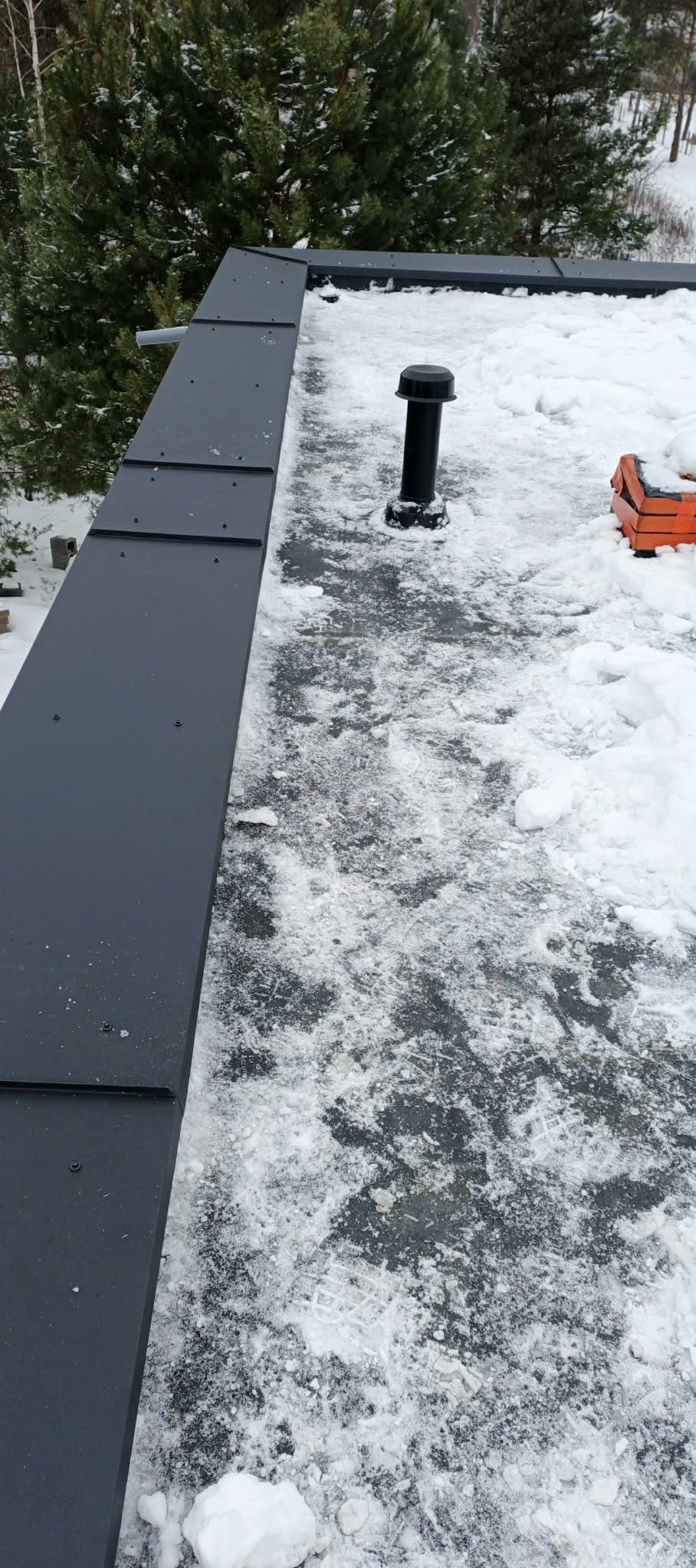 Grafitowy dach pokryty śniegiem, widoczny komin wentylacyjny i fragment skrzynki narzędziowej. Nowoczesne krycie dachu w zimowej scenerii.