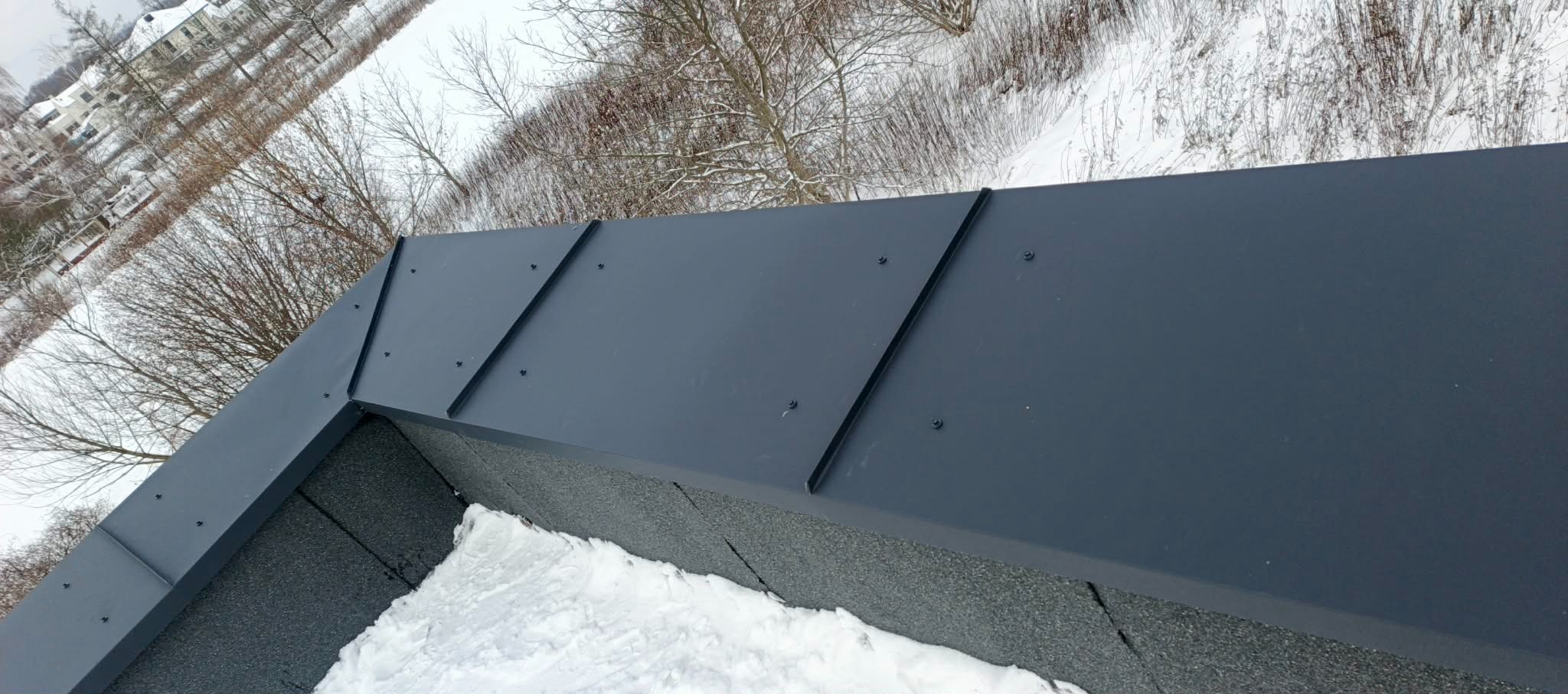Szary, metalowy dach z widocznymi łączeniami blach, pokryty śniegiem. Ujęcie z góry, w tle ośnieżone drzewa i zabudowania.