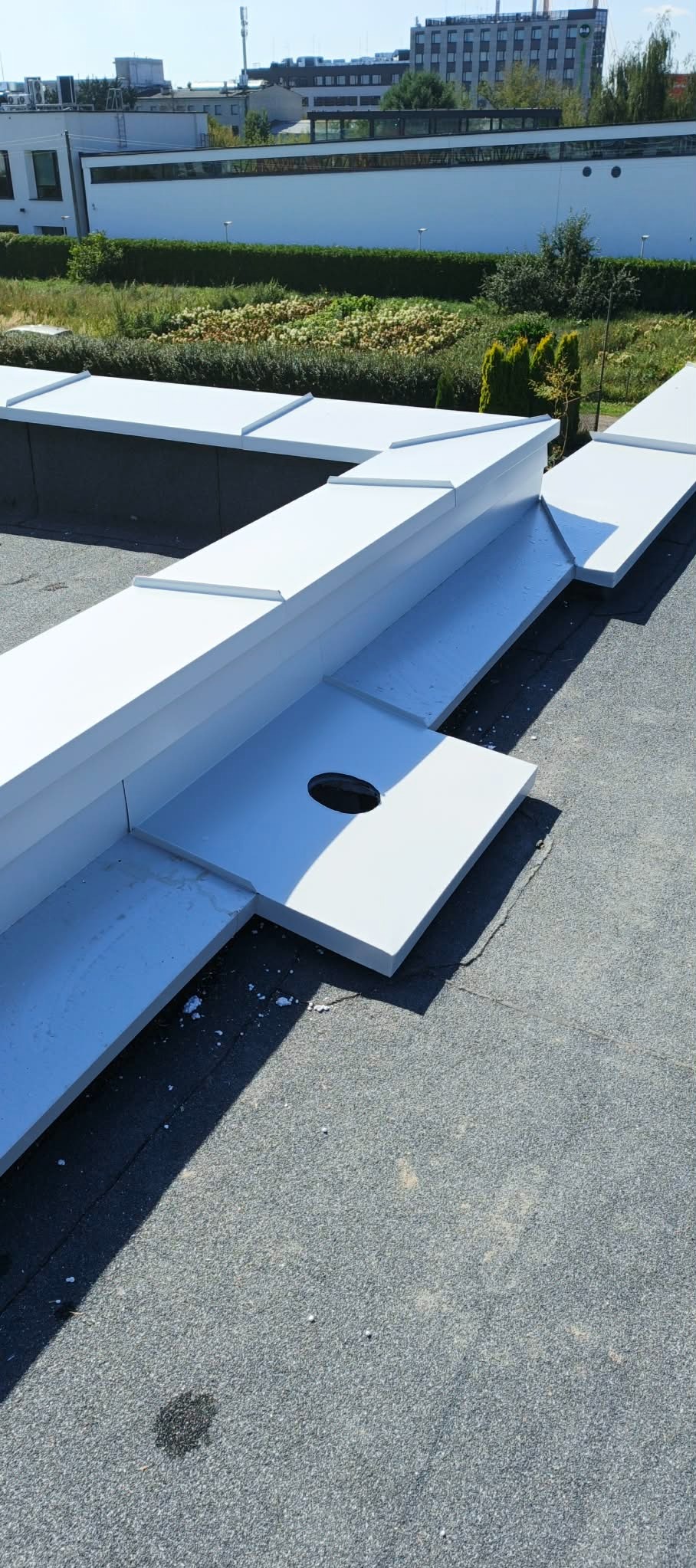 Białe, metalowe obróbki blacharskie na dachu budynku, z widocznym otworem odpływowym. Tło stanowią budynki i zieleń. Ujęcie z góry, w słoneczny dzień.