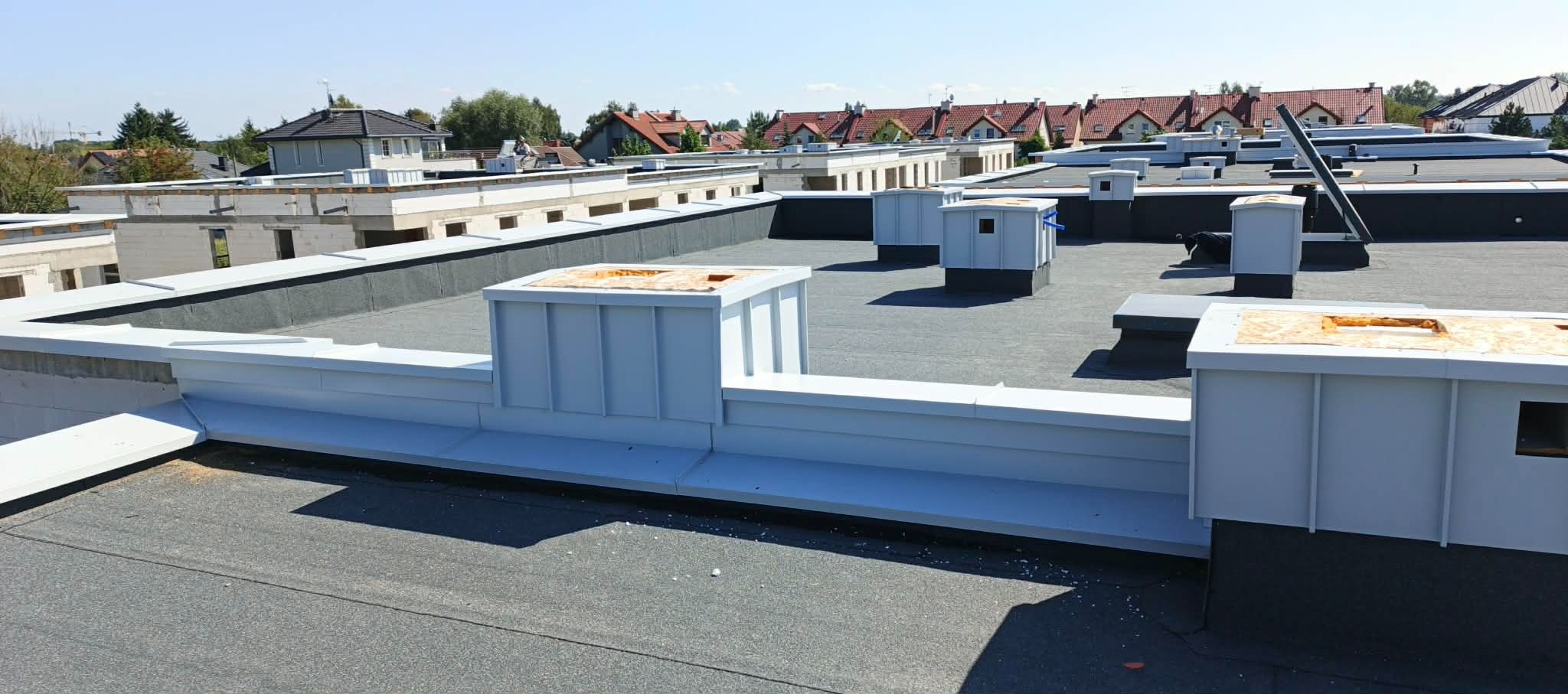 Wykończony płaski dach pokryty papą termozgrzewalną z obróbkami blacharskimi w kolorze białym. Widoczne kominki wentylacyjne i elementy attyki. Tło stanowią budynki mieszkalne.