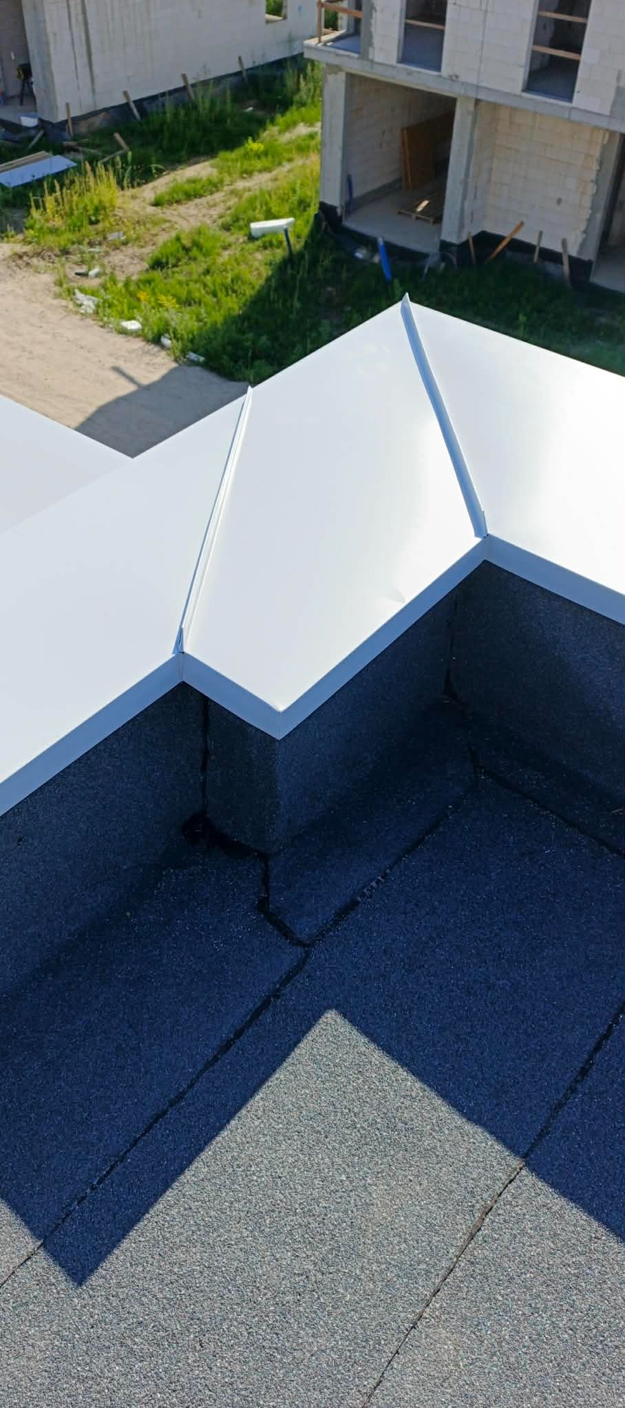 Szczegółowe ujęcie dachu z białymi obróbkami blacharskimi i czarną papą termozgrzewalną na tle surowego budynku w budowie i zieleni. Widok z góry.