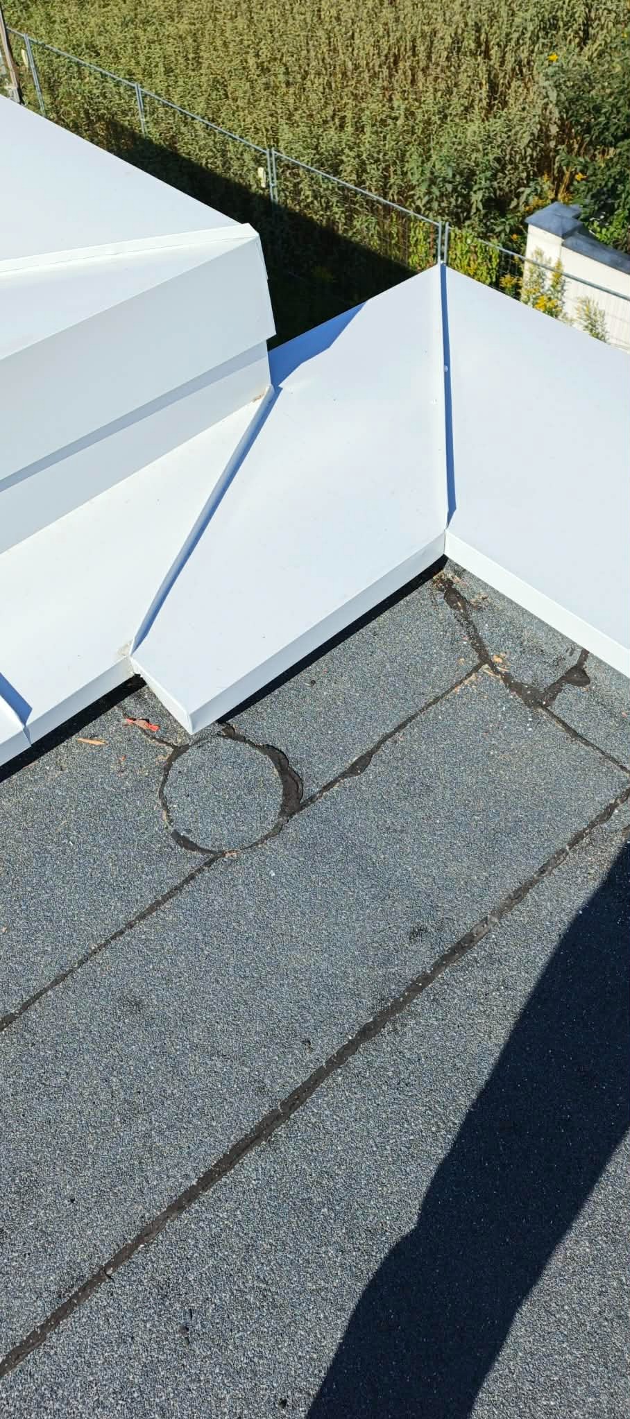 Szczegółowe ujęcie dachu pokrytego papą z widocznymi obróbkami blacharskimi w kolorze białym. Widoczne ślady po uszczelnieniu wokół komina.