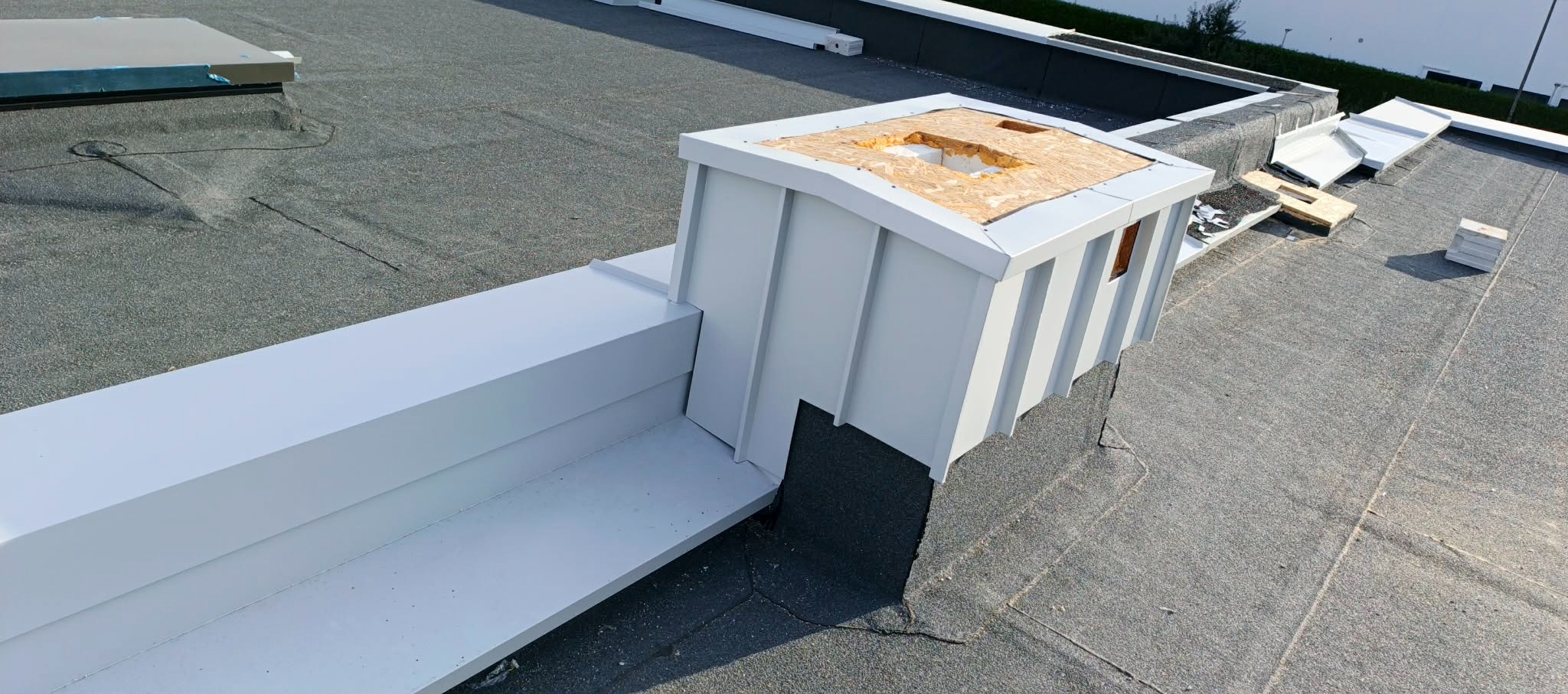 Szczegółowe ujęcie płaskiego dachu z wykończoną obróbką blacharską komina wentylacyjnego, z widocznymi elementami montażowymi i papą dachową.