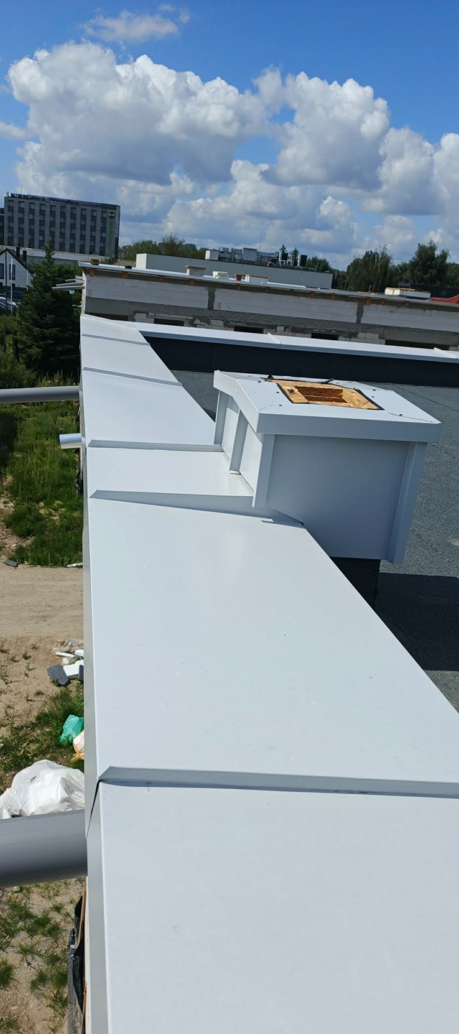 Białe, metalowe obróbki blacharskie na dachu budynku, z widocznym kominem i fragmentem rynny. W tle budynek i błękitne niebo z chmurami.