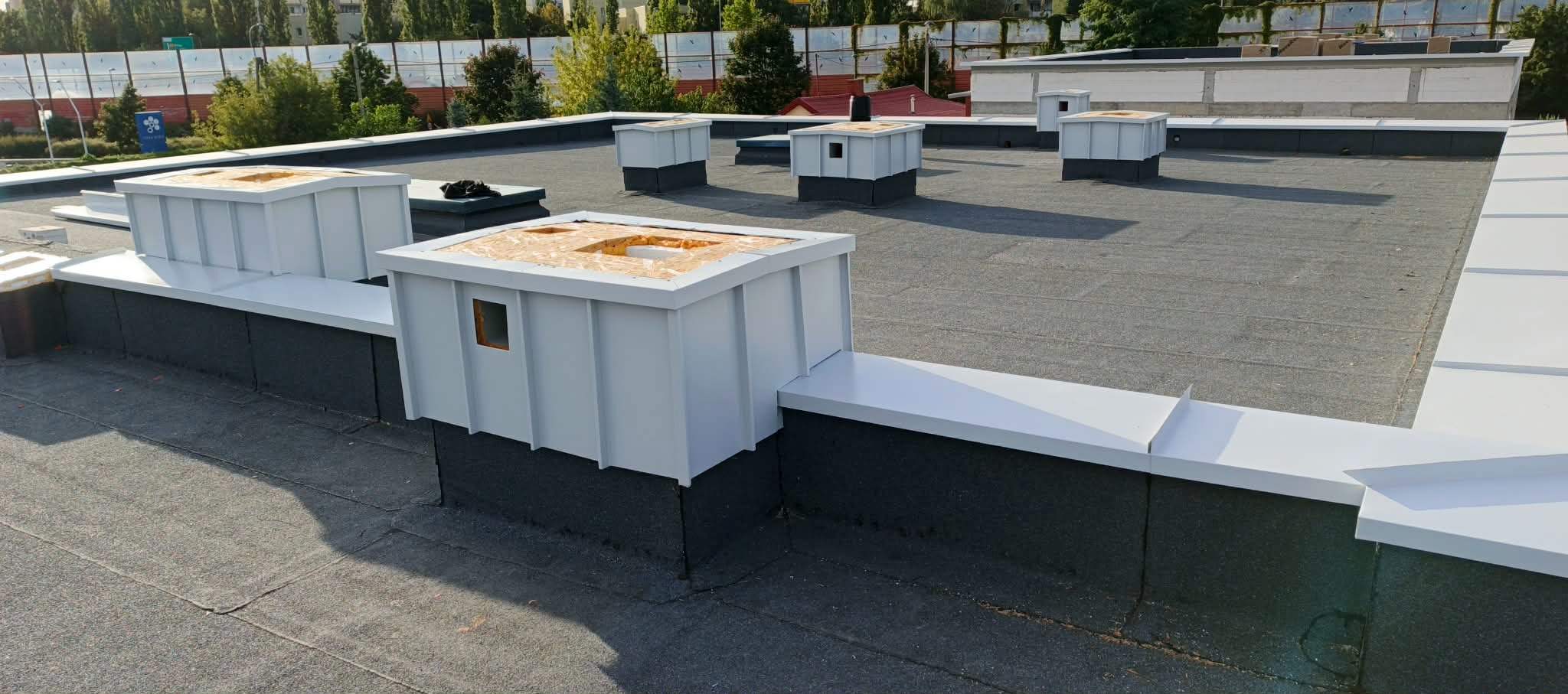 Szary dach pokryty papą termozgrzewalną z obróbkami blacharskimi w kolorze białym, wykończone kominy wentylacyjne. Widok z góry na płaski dach budynku.