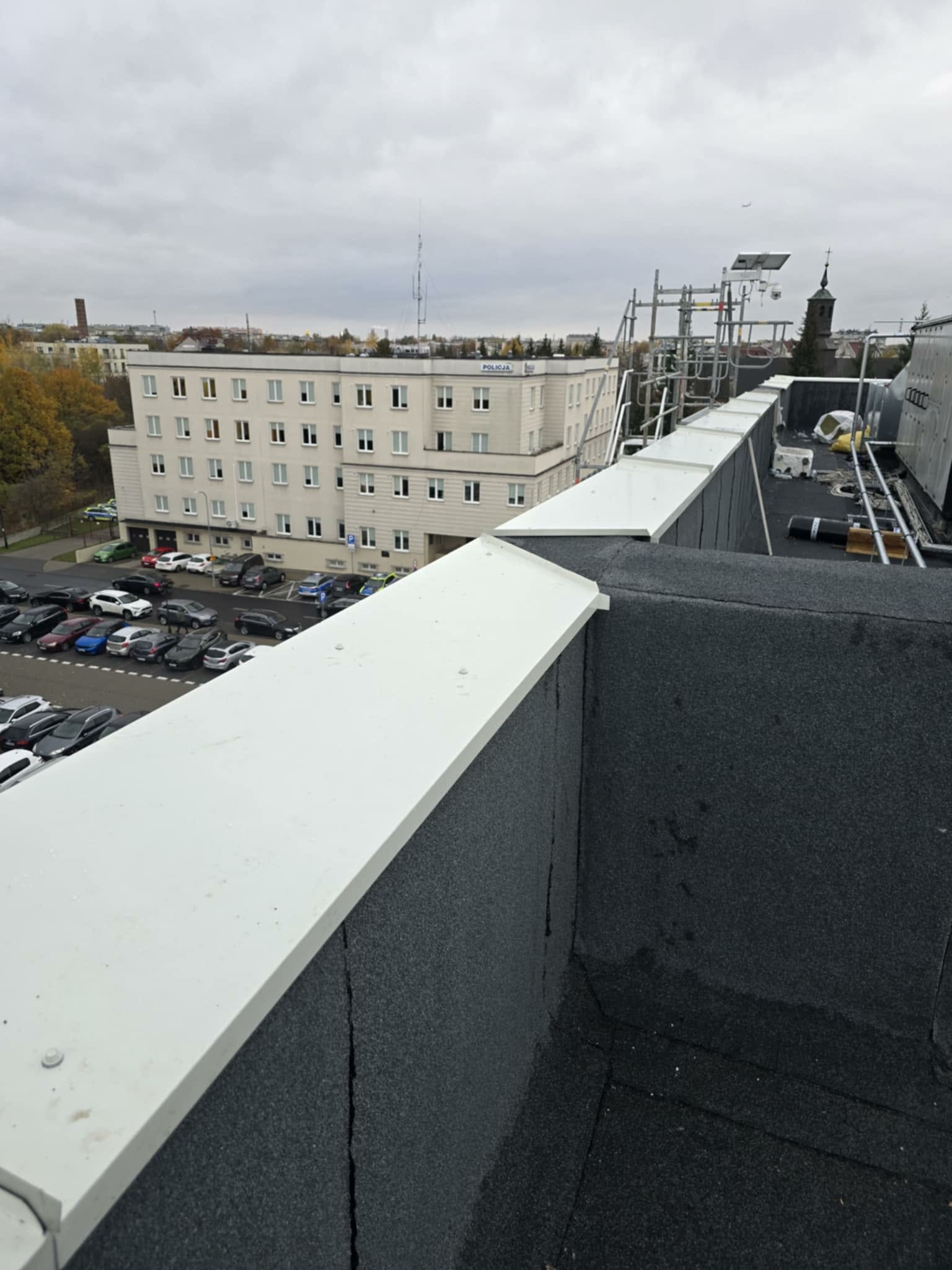 Wykończony fragment dachu pokrytego papą z białymi obróbkami blacharskimi. W tle widoczny budynek i parking.