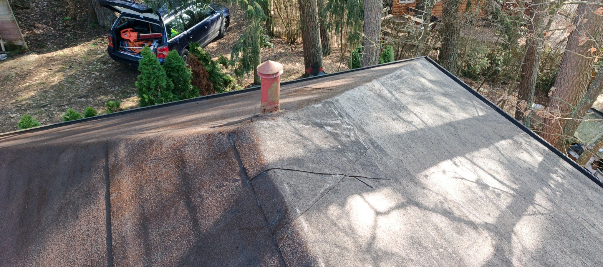 Widok z góry na płaski dach pokryty papą z widocznym kominem wentylacyjnym i szczelinami. W tle zaparkowany samochód z otwartym bagażnikiem i narzędziami.