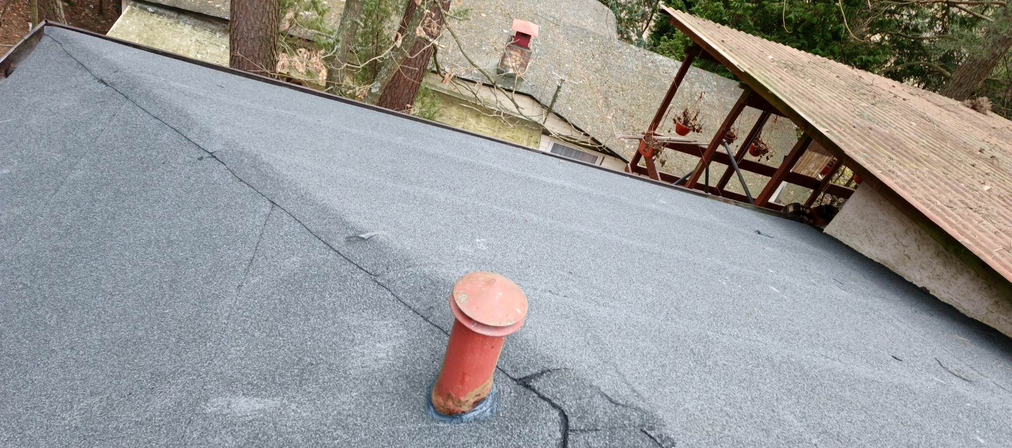 Szary dach pokryty papą z kominkiem wentylacyjnym na tle drzew i fragmentu innego dachu. Widoczne łączenia papy. Ujęcie z góry, lekko z ukosa.