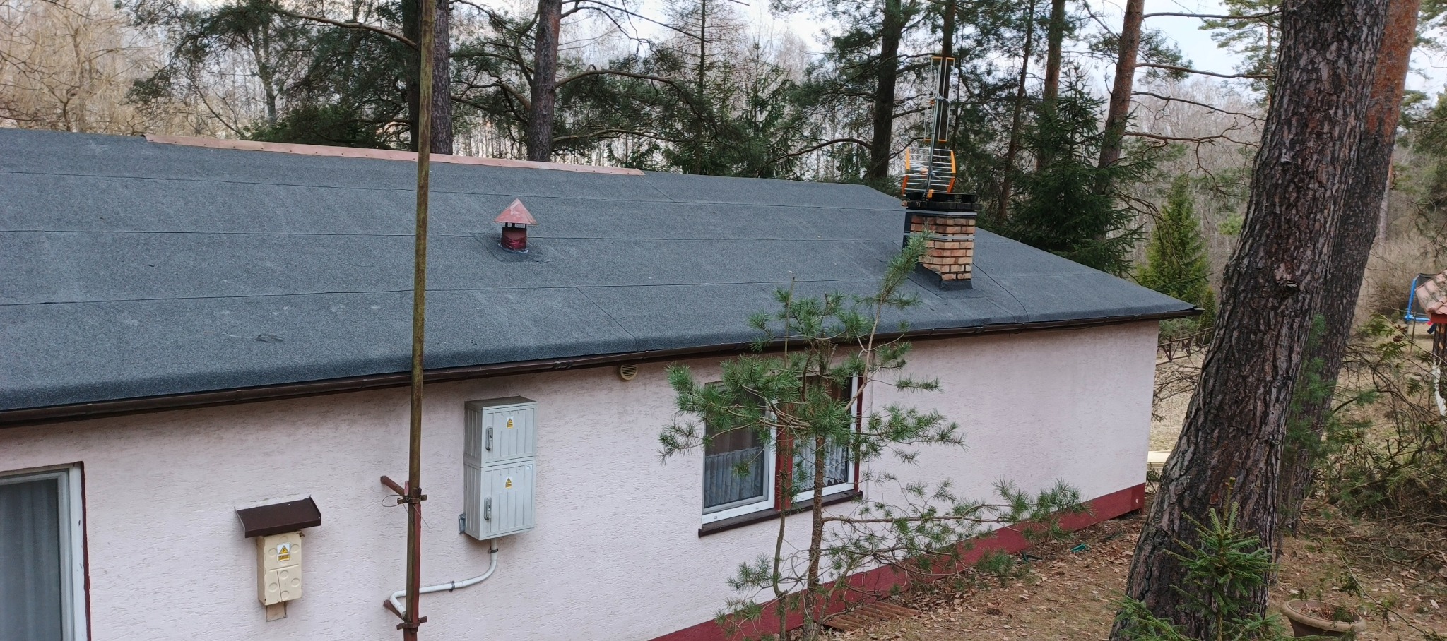 Szary dach z papy termozgrzewalnej na parterowym domu z kominem z cegły, zabezpieczonym metalową konstrukcją. Widok z góry, w tle drzewa.