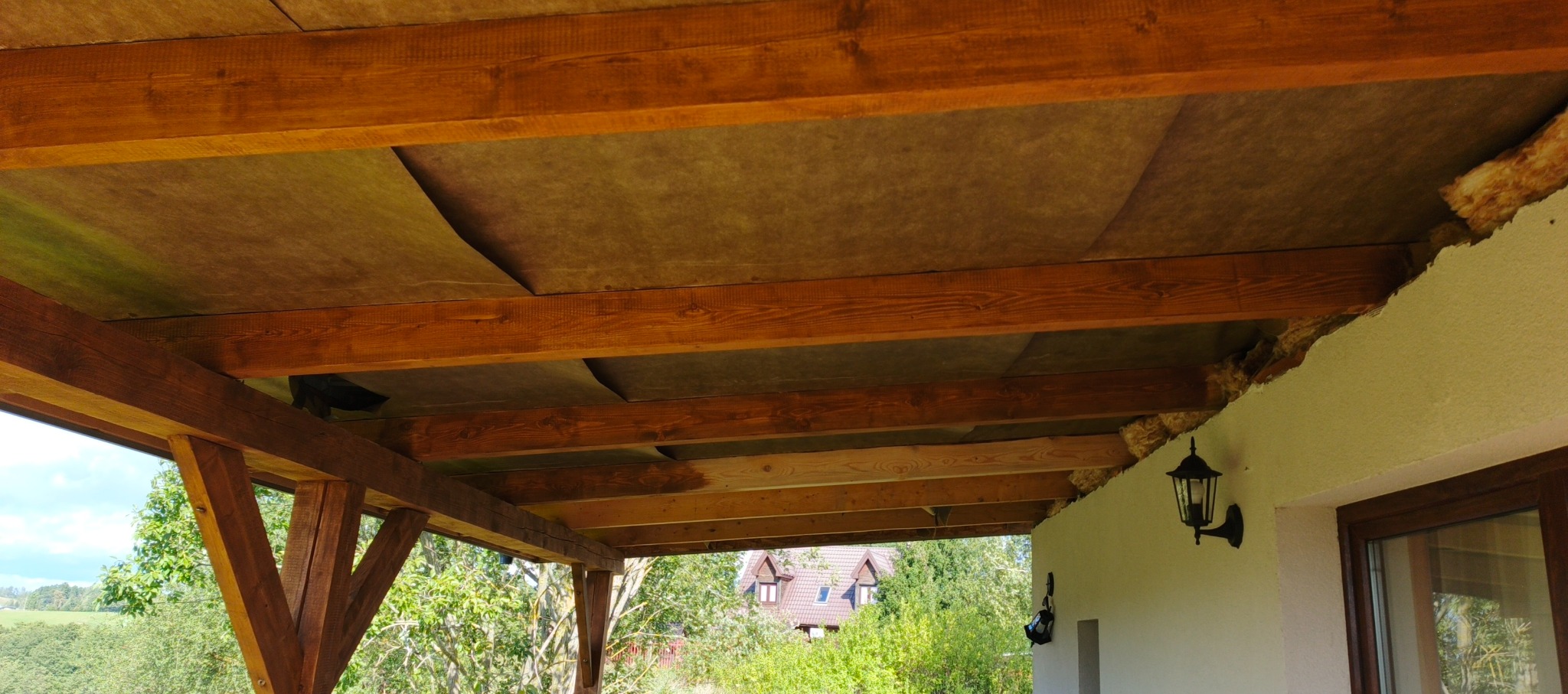 Drewniana konstrukcja dachu z izolacją, widok od dołu. Widoczne brązowe belki, szara izolacja i fragment ściany budynku z lampą. W tle zieleń i dom.