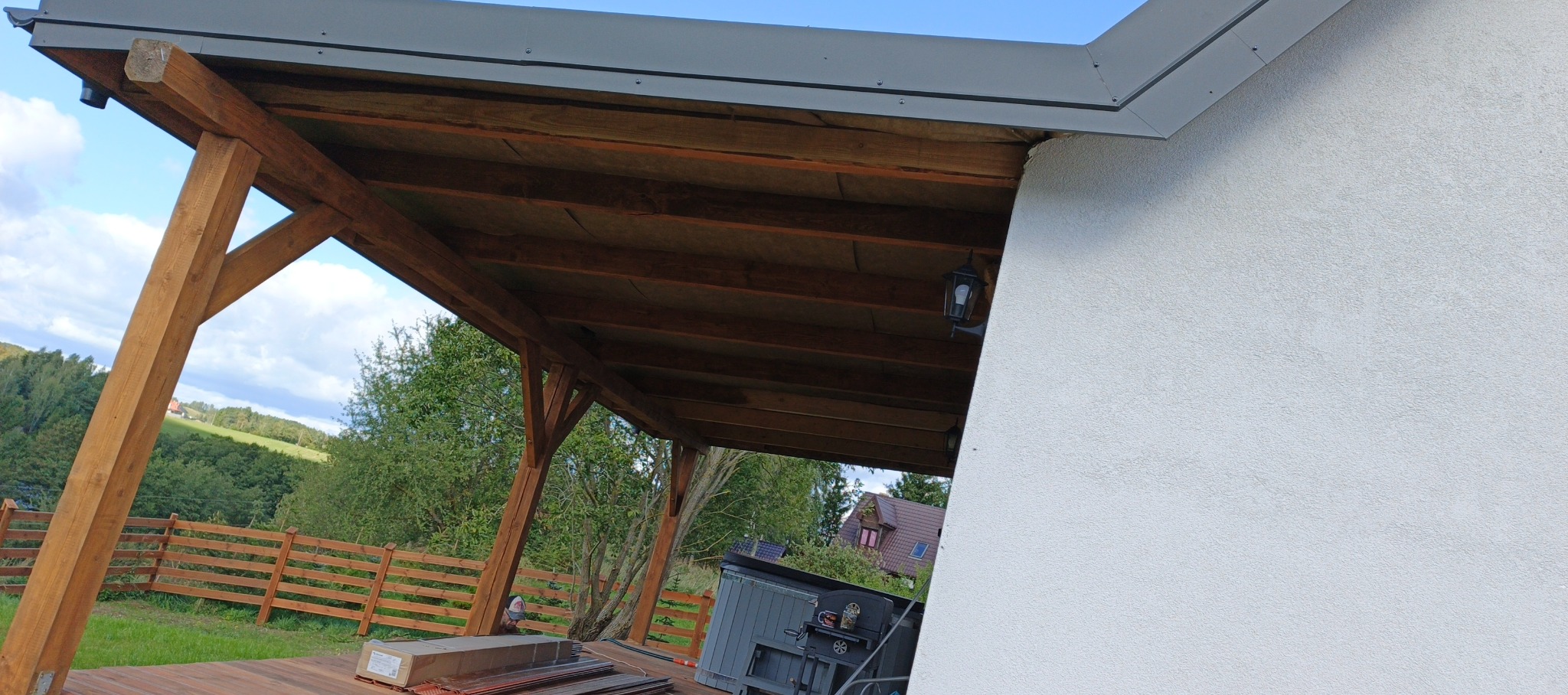 Drewniana konstrukcja zadaszenia tarasu przylegająca do białej ściany budynku. Widoczne elementy dachu, lampa zewnętrzna i fragment krajobrazu.