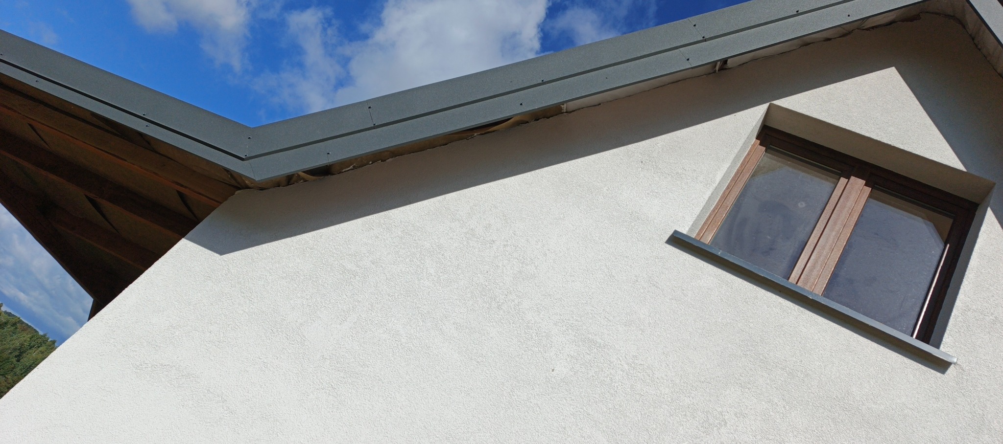 Fragment elewacji domu z oknem i szarą obróbką blacharską dachu na tle błękitnego nieba. Widoczny fragment drewnianej konstrukcji dachu.