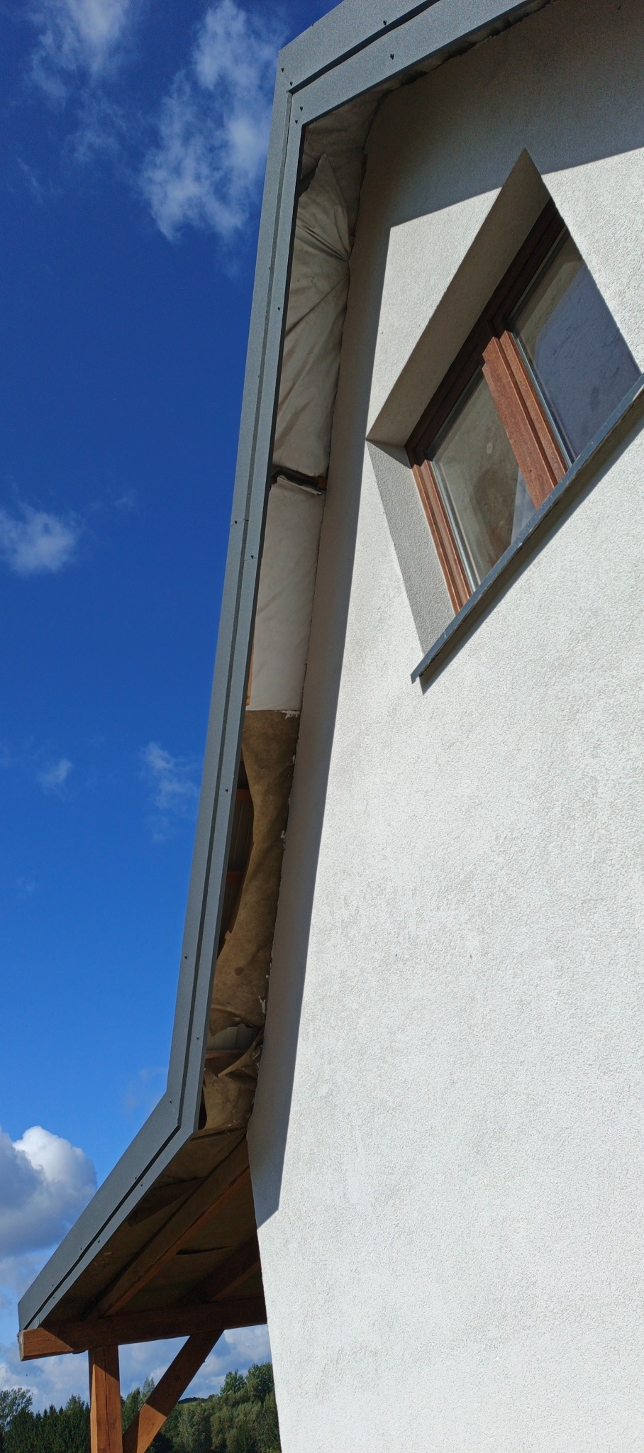 Fragment dachu z szarą obróbką blacharską i izolacją, widok z dołu na tle błękitnego nieba. Detal okna w elewacji budynku. Ujęcie z niskiego kąta.