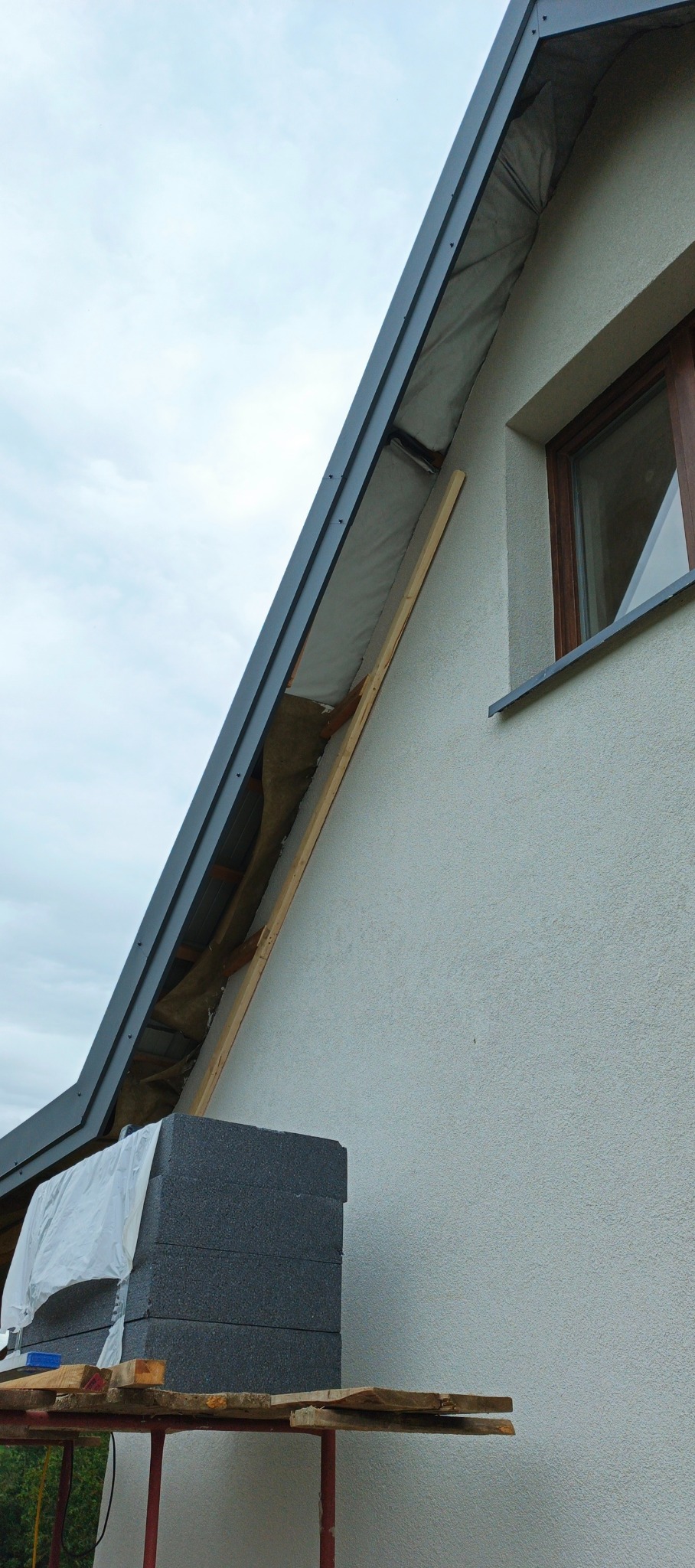 Fragment dachu w trakcie prac. Widoczne ocieplenie, elementy konstrukcyjne i blacha. Na rusztowaniu leżą materiały izolacyjne. Ujęcie z dołu, pochmurne niebo.