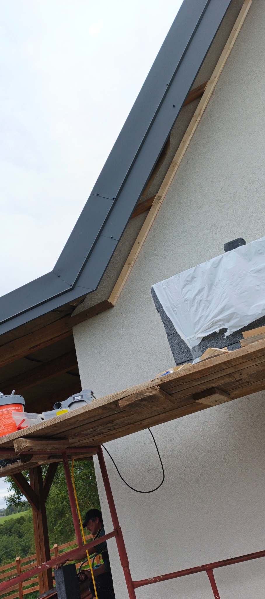 Szary, metalowy element dachu zamontowany na drewnianej konstrukcji, widok z rusztowania. Część ściany otynkowana, fragment ocieplenia zabezpieczony folią.