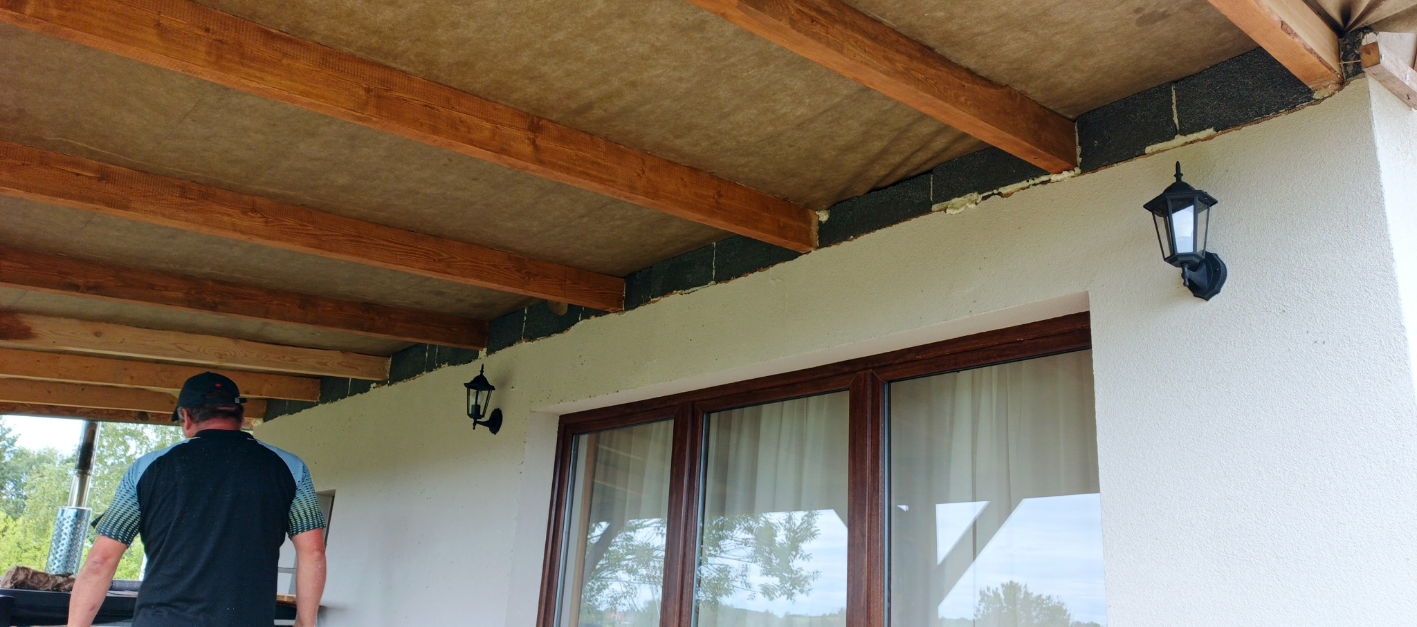 Ocieplenie poddasza wełną mineralną widoczne od zewnątrz, z drewnianą konstrukcją nośną i oświetleniem elewacyjnym. Fragment budynku z oknem i osobą w tle.