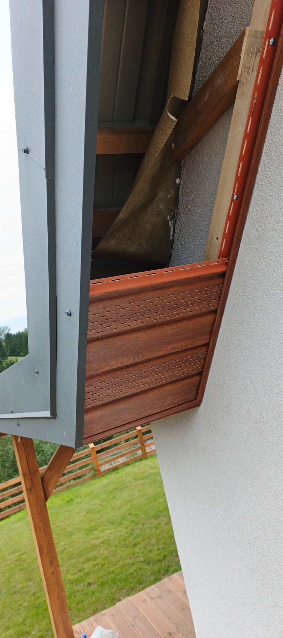 Wykończenie okapu dachu z brązową podbitką drewnopodobną i szarą blachą. Widoczne elementy konstrukcyjne i ocieplenie. Ujęcie z dołu, na tle zieleni.