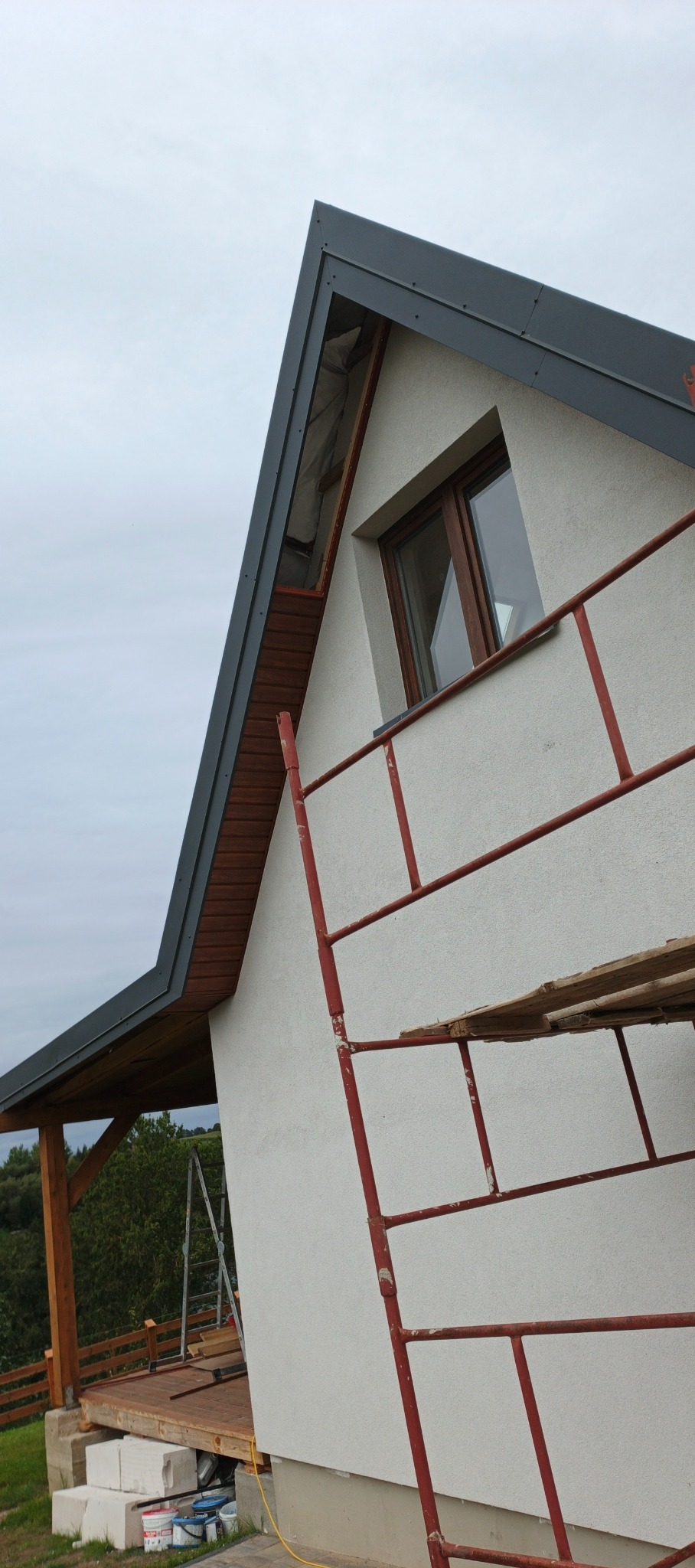 Fragment domu z nowym, grafitowym dachem i drewnianą podbitką, widziany z dołu. Na ścianie rusztowanie. Widoczne okno i fragment tarasu z drewnianą konstrukcją.