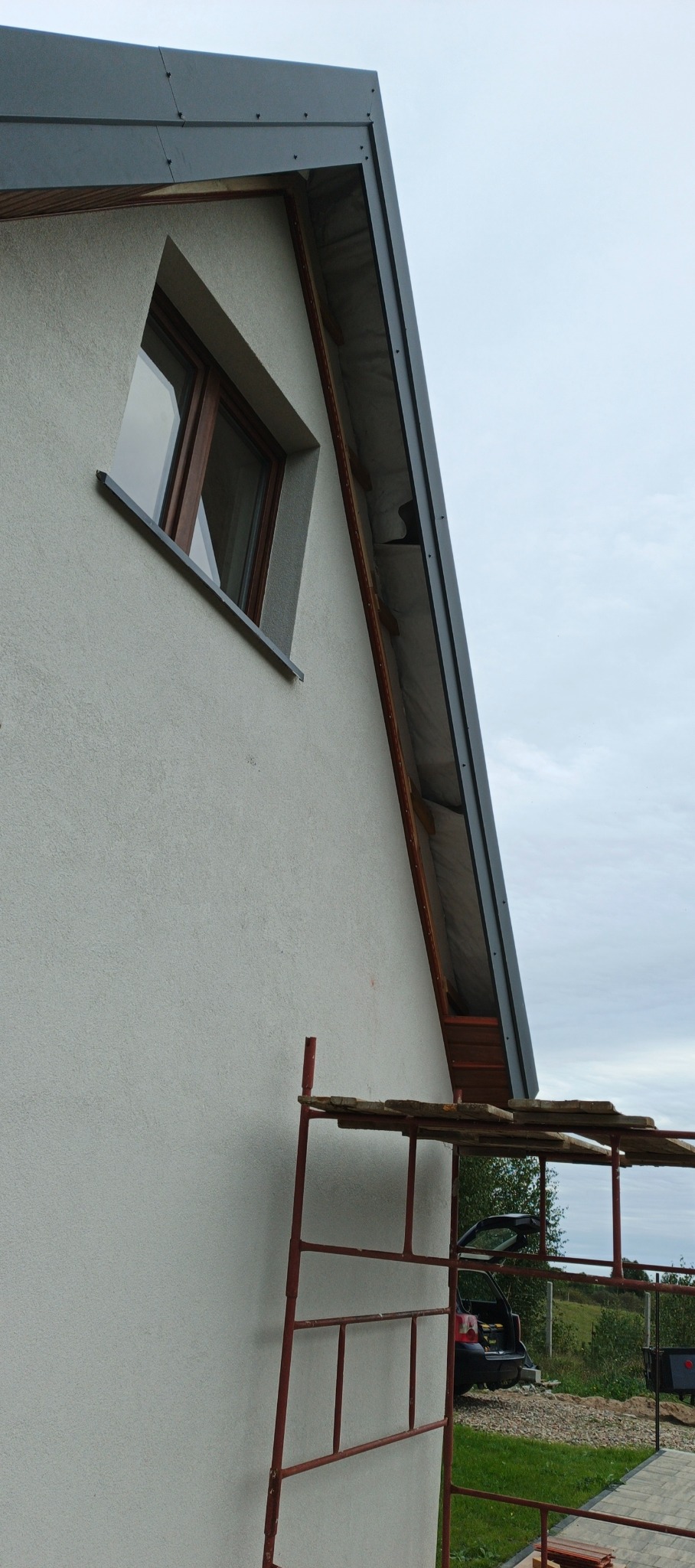 Fragment ściany domu z oknem i fragmentem dachu z szarą obróbką blacharską. Widoczne rusztowanie. Ujęcie z dołu, pochmurne niebo w tle.