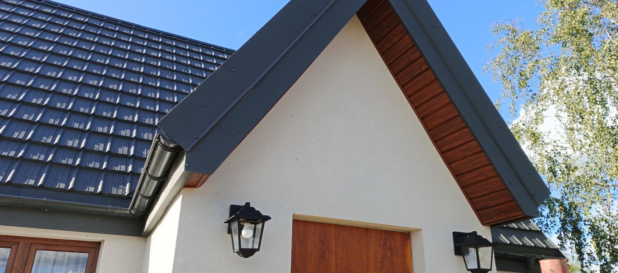 Fragment domu z ciemną dachówką, rynnami i obróbkami blacharskimi. Widoczne oświetlenie zewnętrzne i fragment drzewa na tle błękitnego nieba.