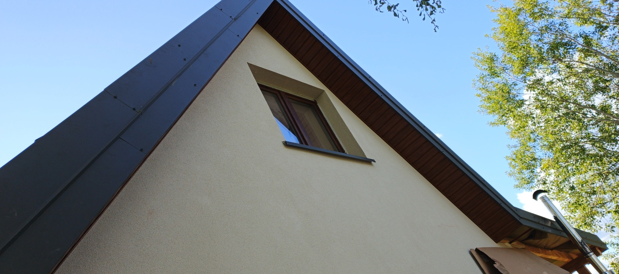 Szary dach z blachy na domu z oknem w kształcie trójkąta, widziany z dołu pod kątem. Na tle błękitne niebo i korona drzewa. Widoczny komin i fragment obróbki blacharskiej.
