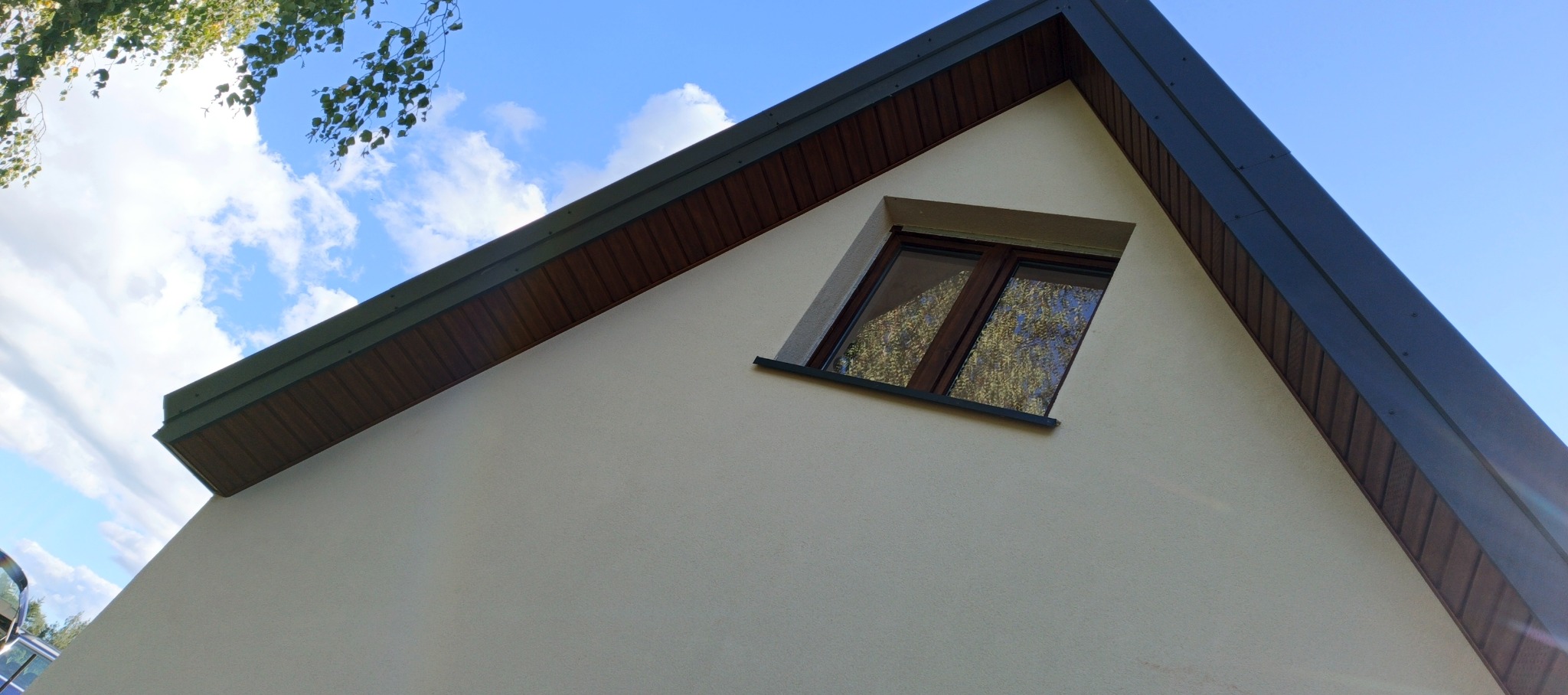 Fragment dachu z oknem poddaszowym, widok z dołu. Ciemna blacha na krawędzi dachu kontrastuje z jasną elewacją. W oknie odbija się zieleń drzew.