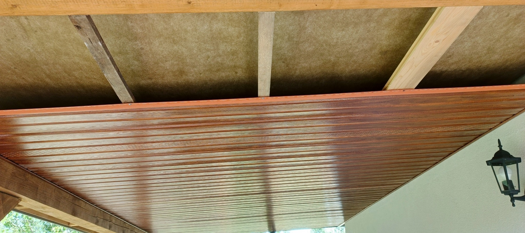 Drewniana podbitka dachowa z widoczną izolacją i elementami konstrukcyjnymi. Ujęcie z dołu, widoczna lampa zewnętrzna.