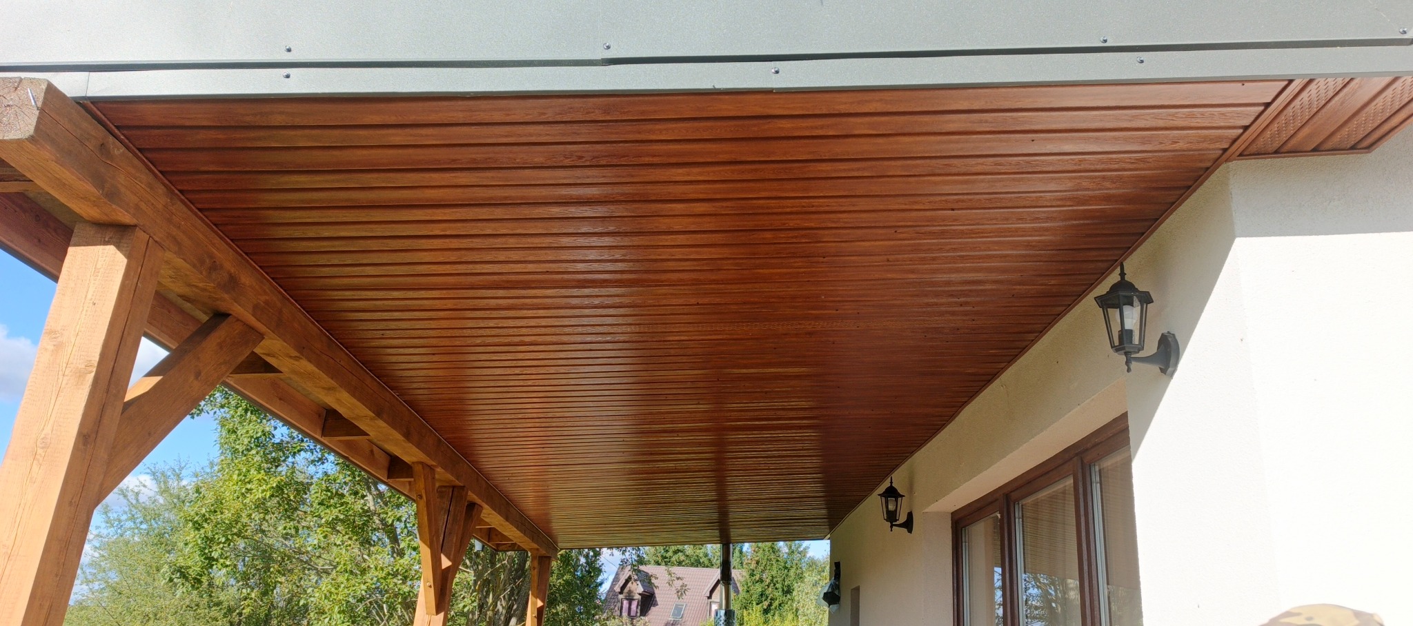 Drewniana podbitka dachowa z widoczną konstrukcją wsporczą i oświetleniem zewnętrznym. Detal wykończenia dachu z bliska, w ciepłej tonacji.