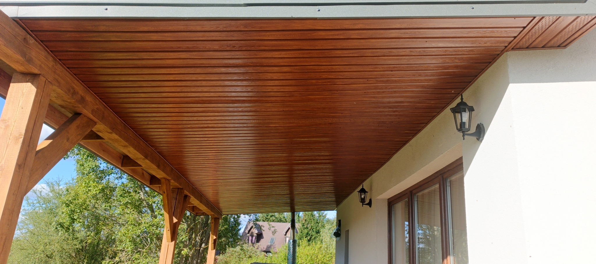 Drewniana podbitka dachowa z widoczną konstrukcją wsporczą i oświetleniem zewnętrznym. Detal wykończenia dachu z bliska, z elementami architektury budynku.