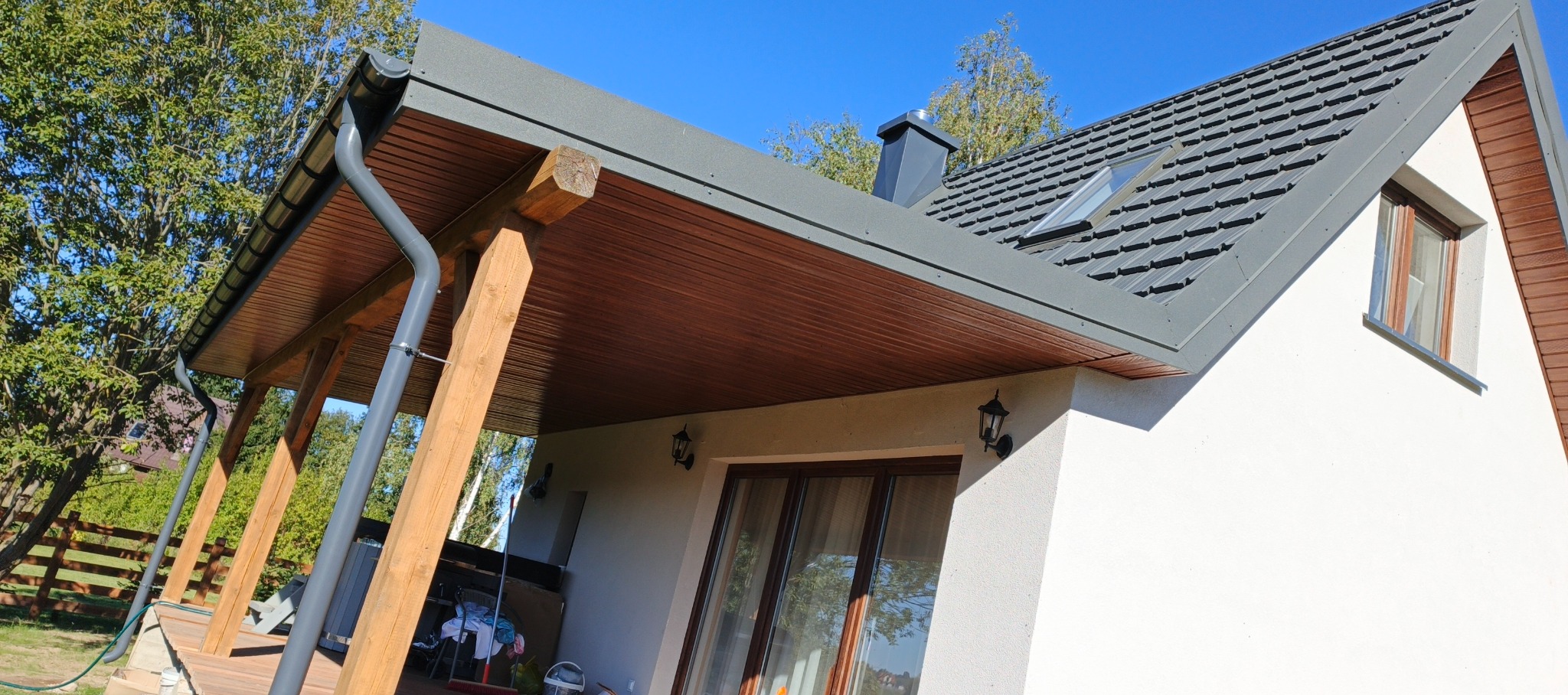 Szary dach z blachodachówki z oknem dachowym i kominem. Widoczne rynny, drewniana podbitka i elementy konstrukcyjne z drewna. Biała elewacja budynku.