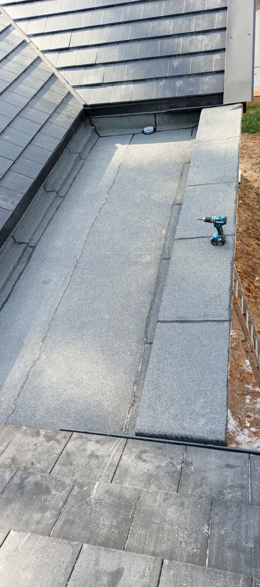 Widok z góry na fragment dachu z papy termozgrzewalnej, na którym leży wkrętarka akumulatorowa. W tle dachówka i drabina oparta o ścianę budynku.