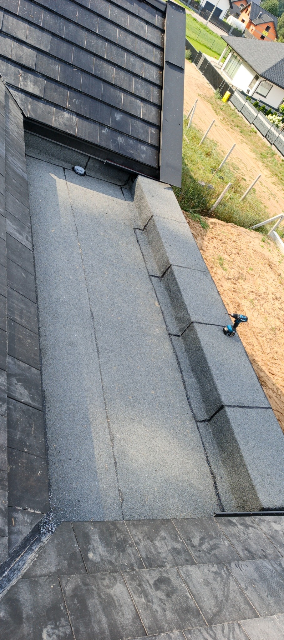 Fragment dachu z papy termozgrzewalnej, widoczny fragment orynnowania i narzędzie pracy - wkrętarka. Ujęcie z góry, w tle zabudowa jednorodzinna.