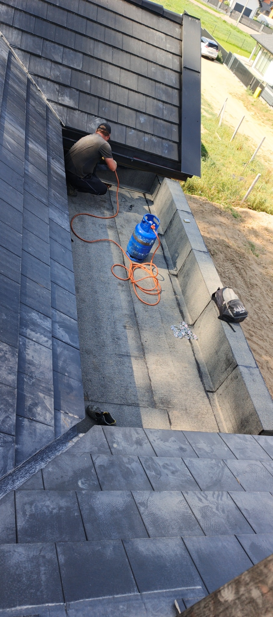 Dekarz pracuje na dachu z czarnej dachówki, używając palnika podłączonego do niebieskiej butli gazowej. Widoczne narzędzia i elementy konstrukcji dachu.