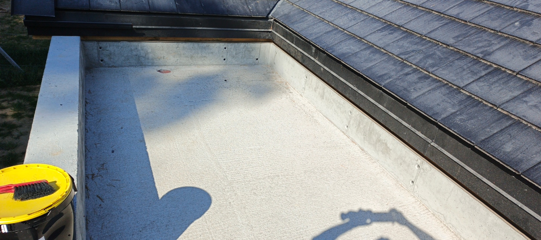 Betonowa attyka dachu z fragmentem pokrycia z dachówki łupkowej. Widoczny żółty pojemnik z pędzlem. Ujęcie z góry, w słoneczny dzień.