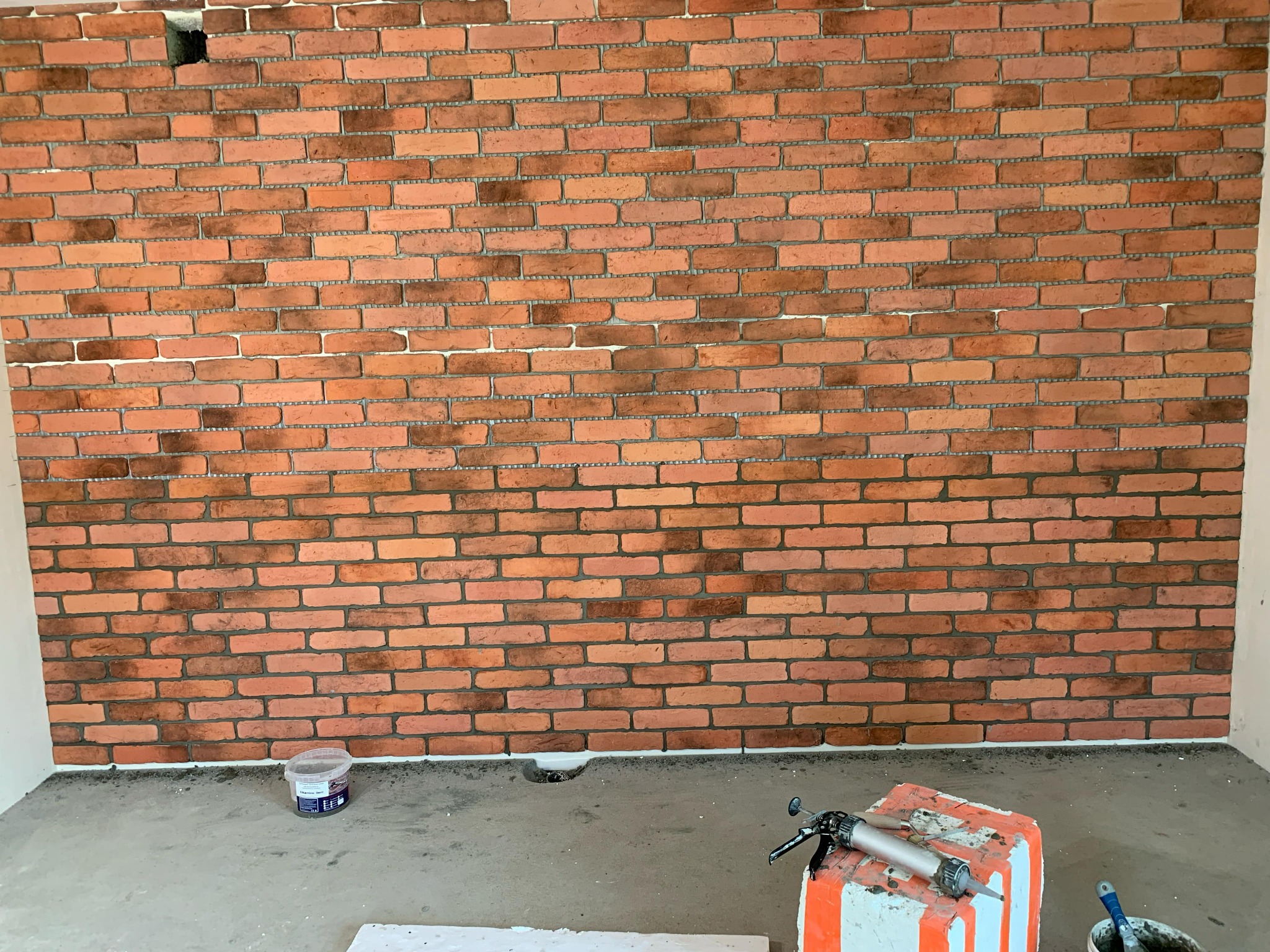 Ściana wyłożona dekoracyjną czerwoną cegłą, z widocznym otworem w górnym rogu i narzędziami glazurniczymi na podłodze, w trakcie prac wykończeniowych.