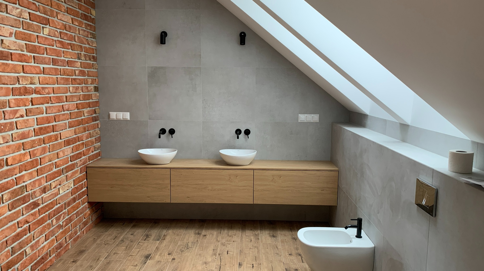 Nowoczesna łazienka na poddaszu z ceglaną ścianą, drewnianą szafką pod umywalki nablatowe, szarą płytką na ścianach i drewnopodobną podłogą, oświetlona światłem wpadającym przez okno dachowe.