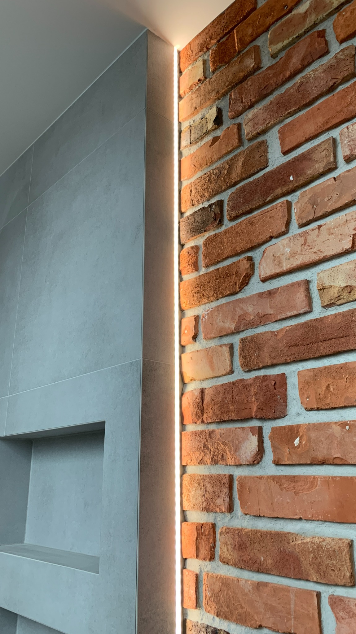 Ściana z czerwonej cegły i szarych płytek z wnęką, oświetlona pionową listwą LED.